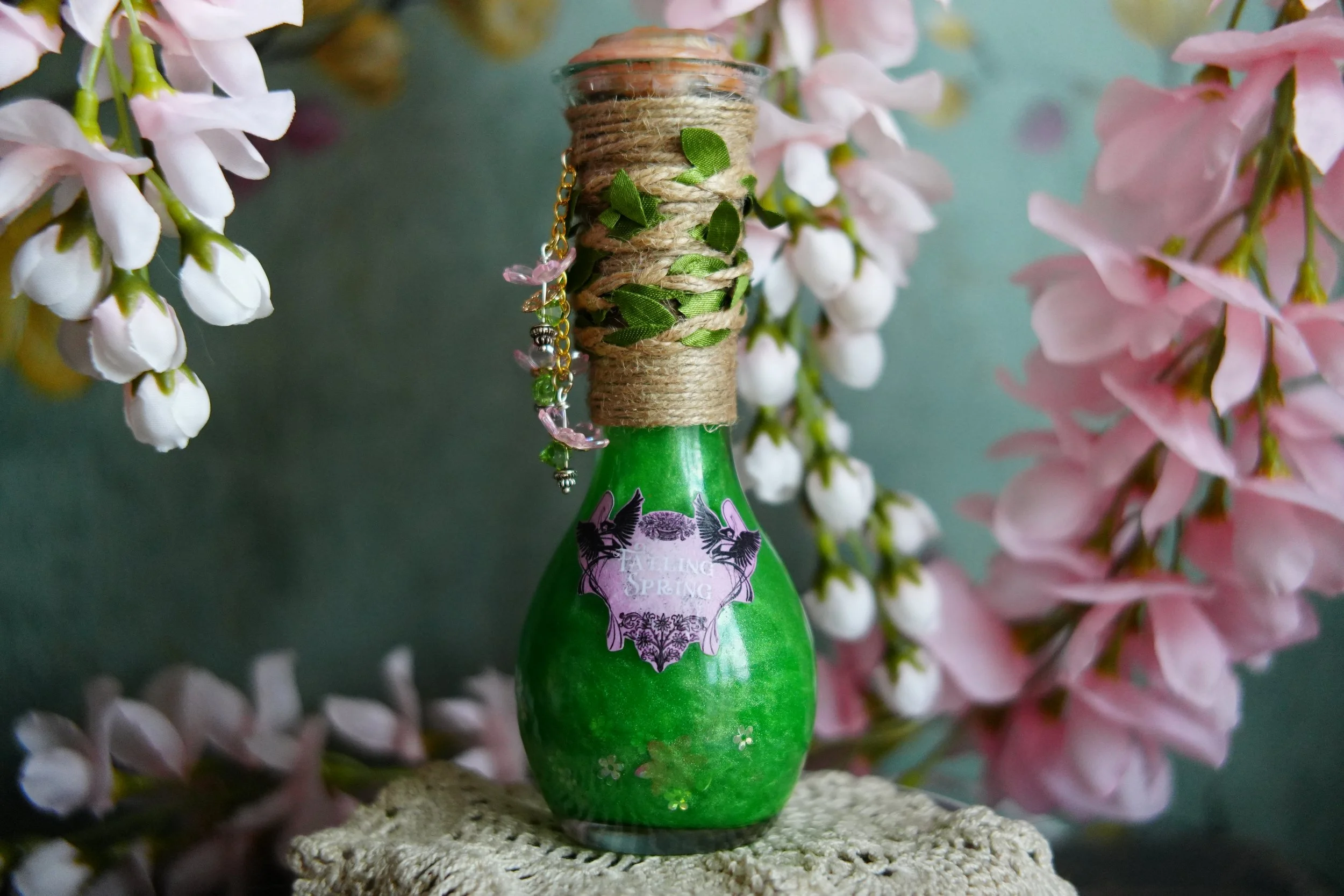 Green decorative potion bottle labeled "Faeling Spring" with twine, green leaves, pink bead charm, and cork, displayed among soft pink blossoms.