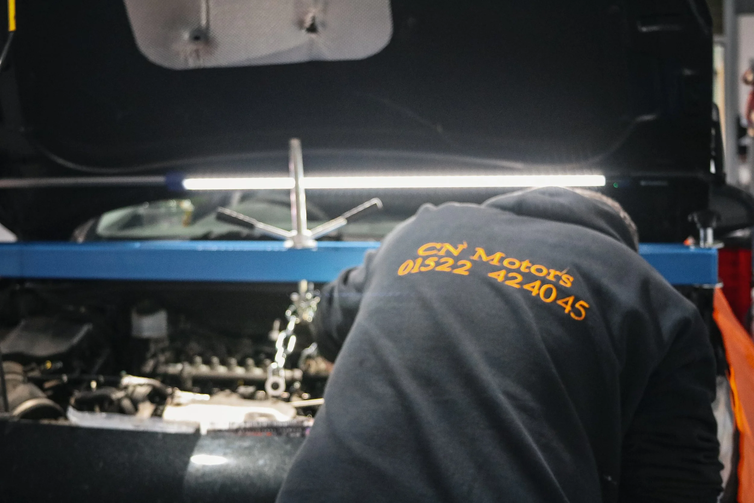 A mechanic working under the open hood of a car in a garage, wearing a black jacket with orange text that reads 'CN Motors' and a phone number.