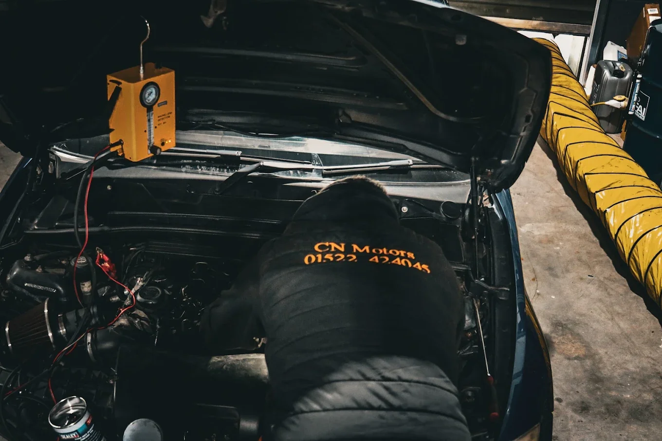 A person working on a car engine in a garage, with the car's hood open and a yellow and black ventilation duct nearby.