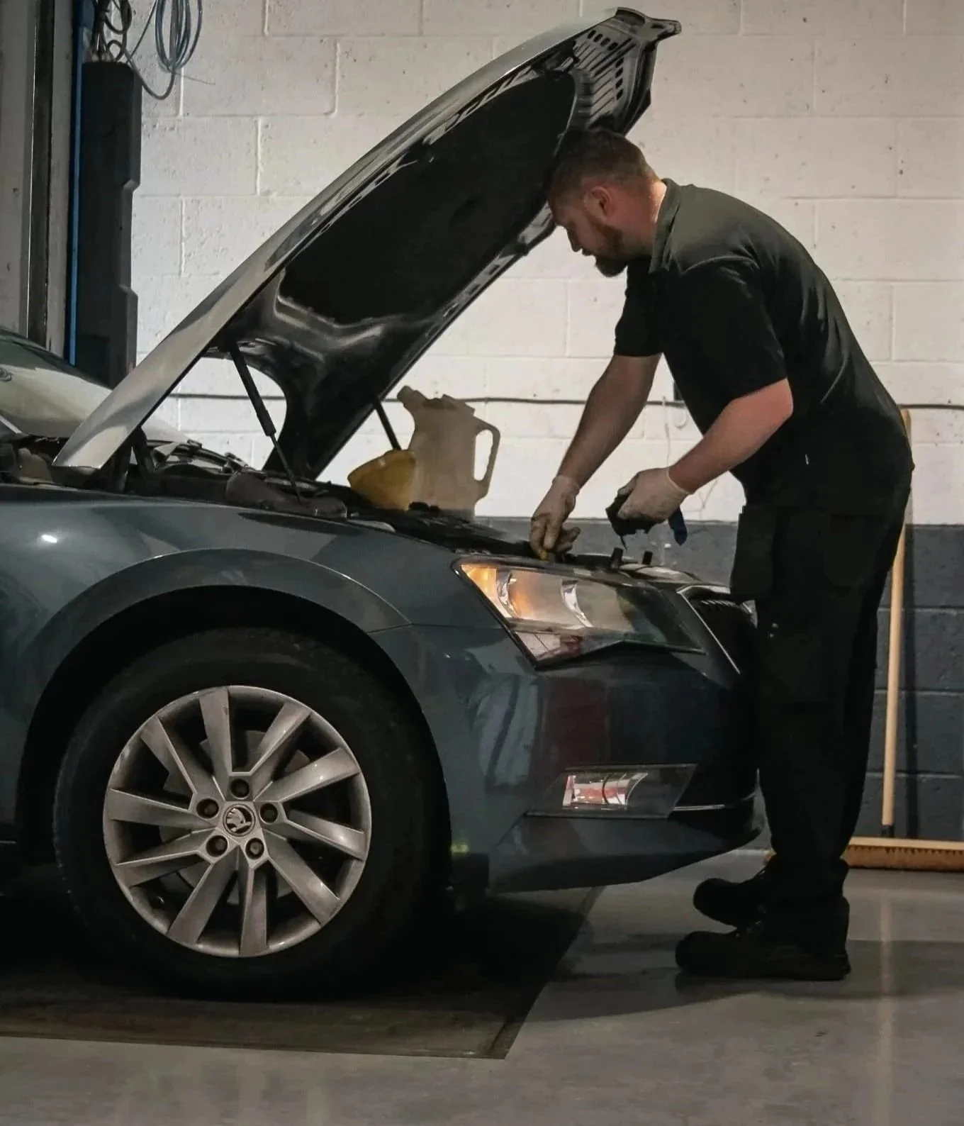 A mechanic working under the hood of a gray car in a garage.