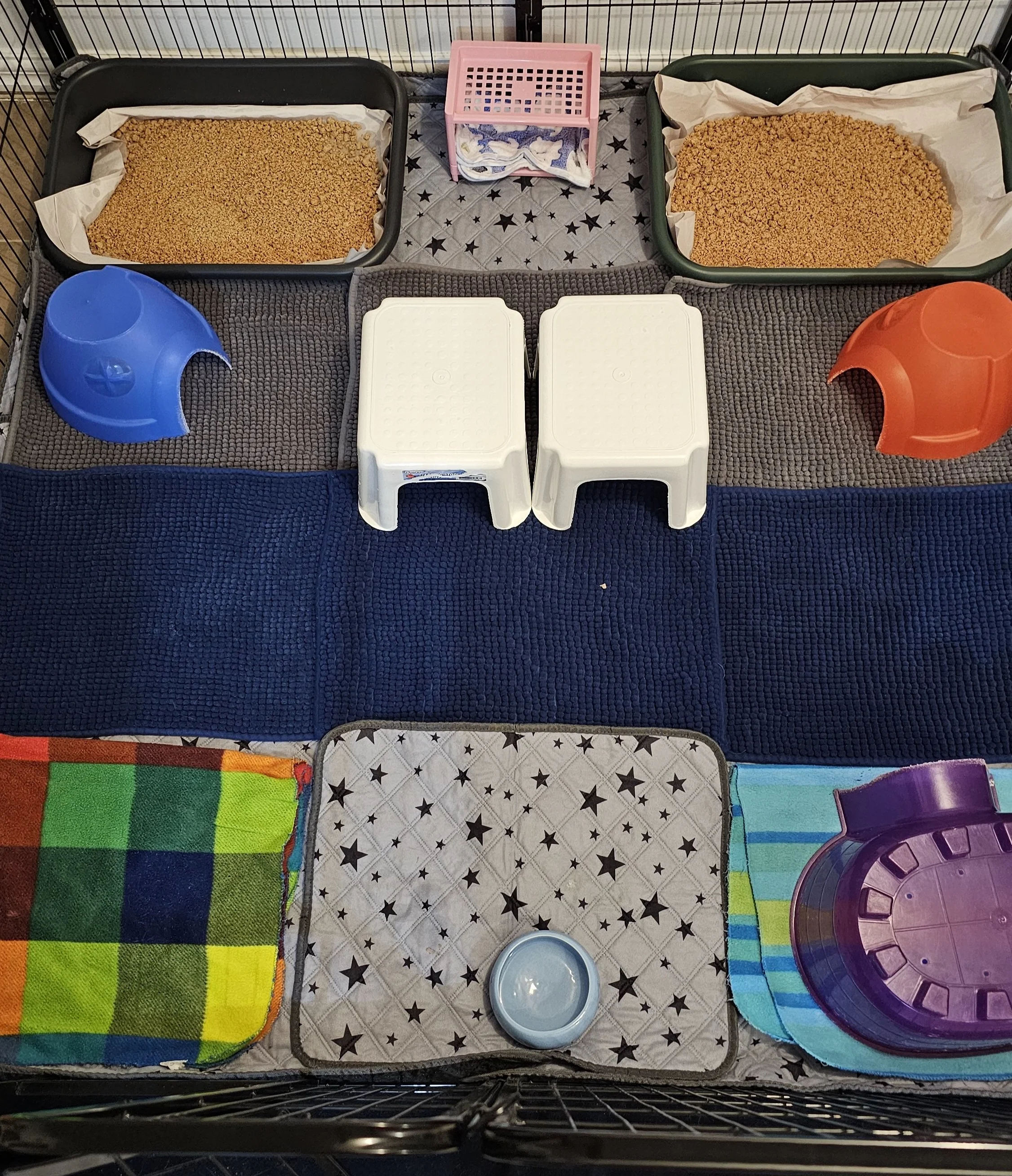 A pet cage setup with two trays of dry kibble food, colorful bowls, small tables, and blankets.