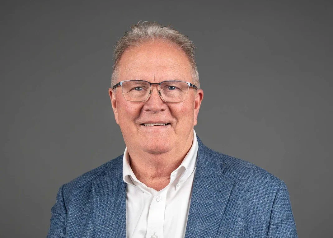Portrait of a middle-aged man with glasses, wearing a blue blazer and white shirt, smiling against a gray background.