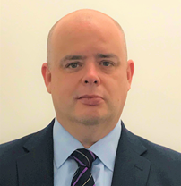 Portrait of a bald man in a business suit and tie, standing against a plain light-colored background.