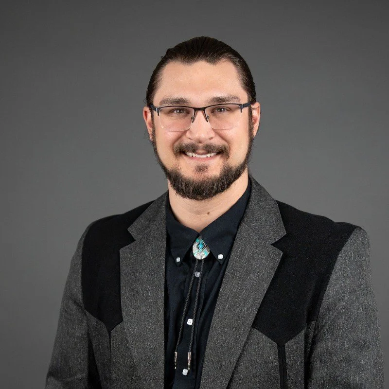 A man with glasses, a beard, and long hair tied back, wearing a dark shirt with a bolo tie, and a gray blazer with black accents, smiling against a plain gray background.