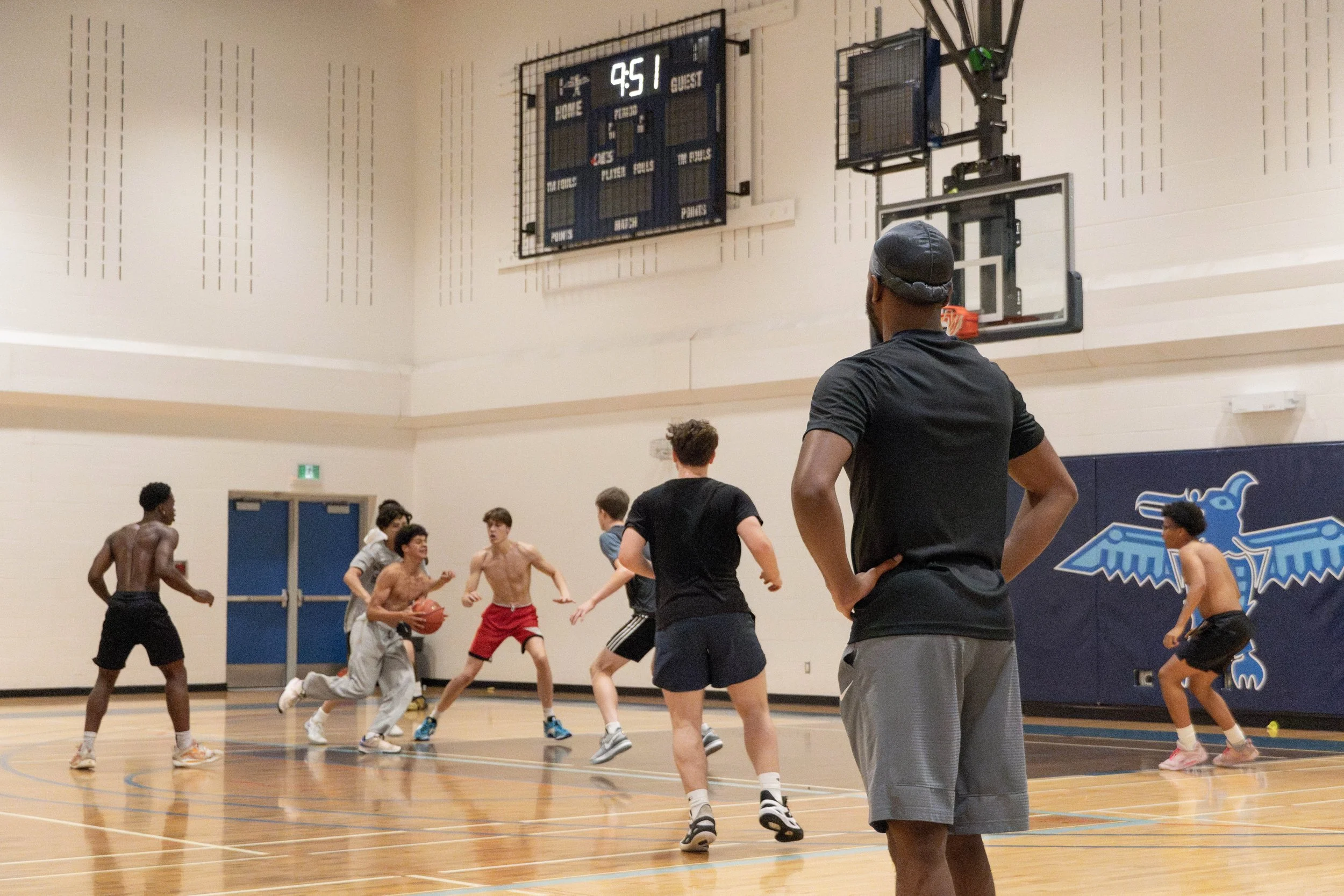 Group of young men playing basketball in an indoor gymnasium, with one man observing from the side. The scoreboard shows 9:51 remaining in the game.
