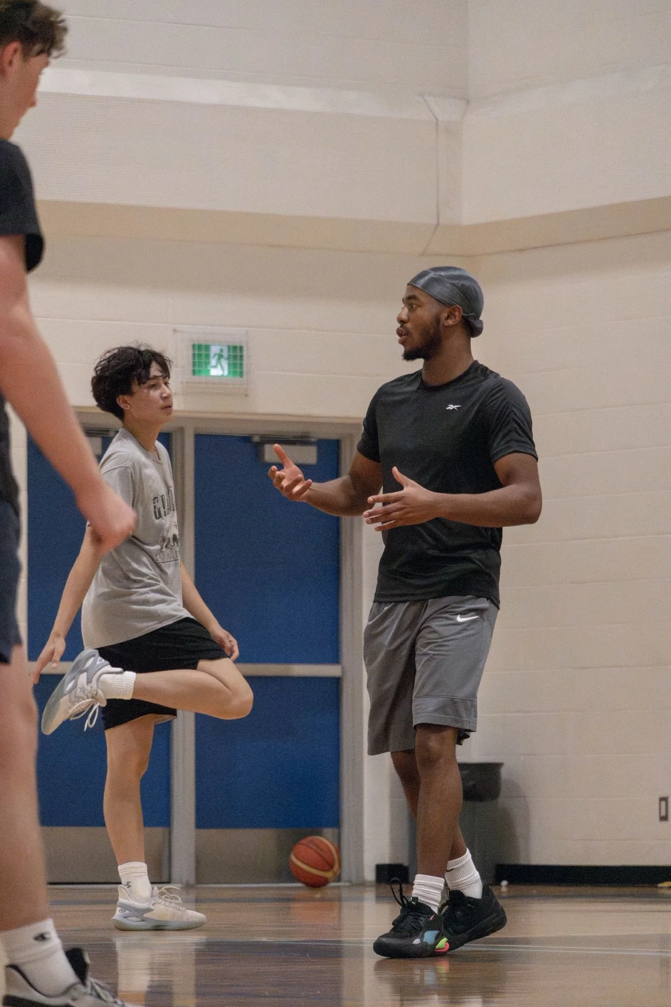 A basketball coach talking to young players in a gymnasium. One girl is stretching her leg while listening, with a basketball on the floor nearby.