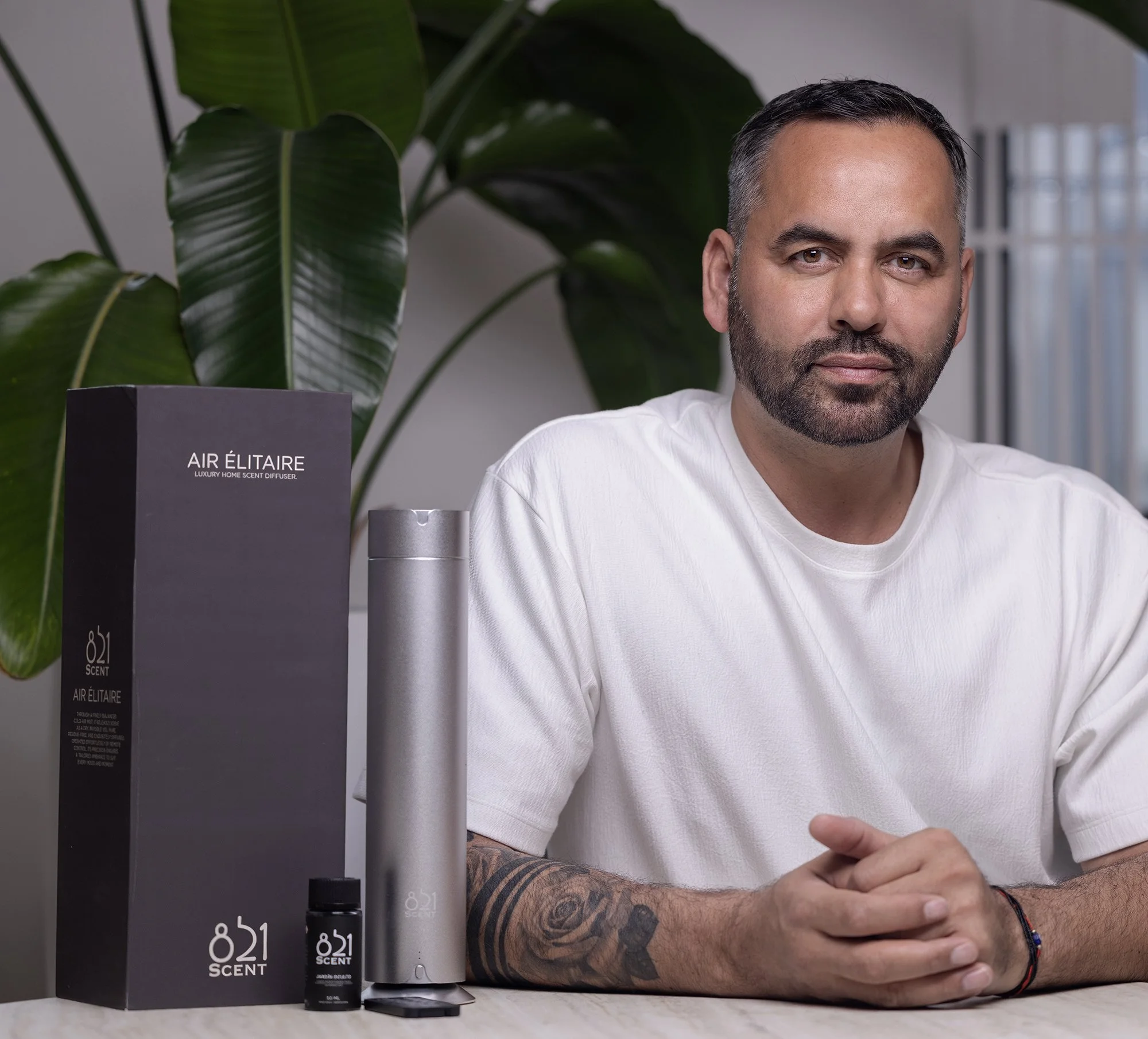A man with a beard and tattoos sitting at a table with a luxury home scent diffuser and a box and bottle of 821 Scent Air Éliteire on display, indoors with a large plant and window in the background.