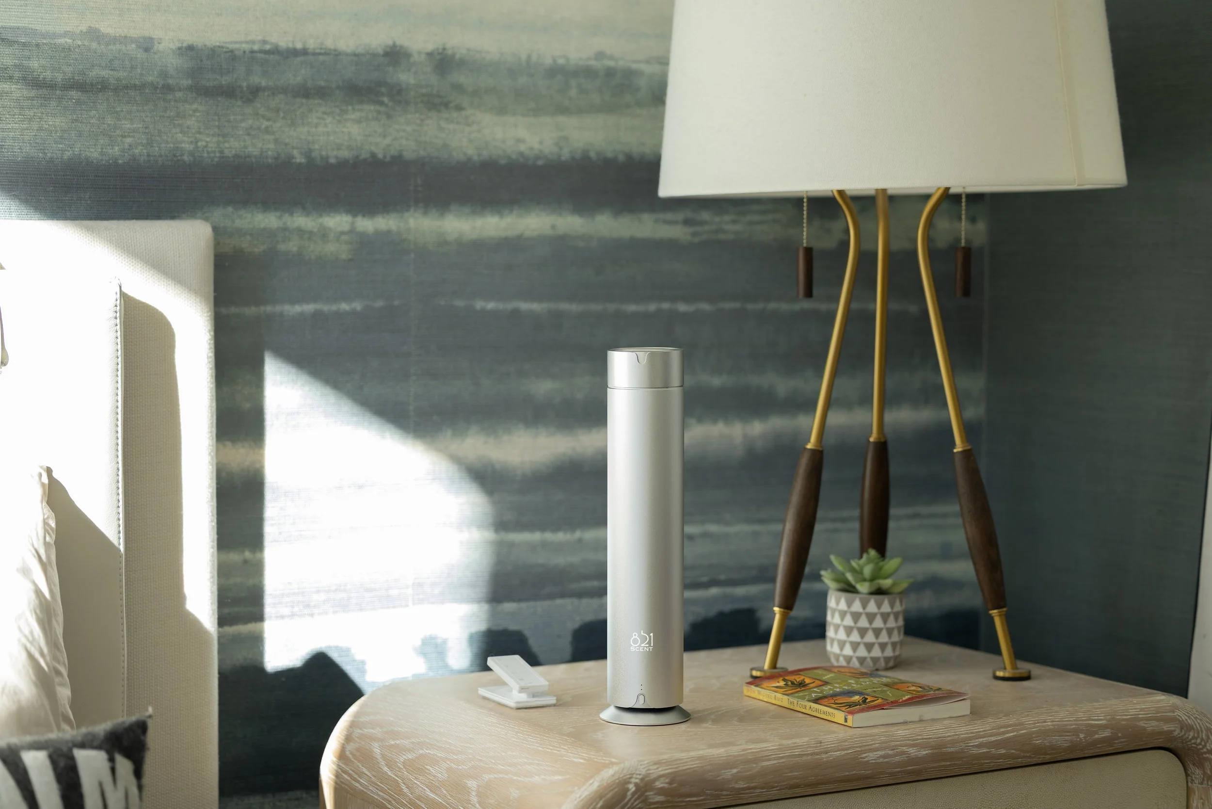 A modern bedroom nightstand holding a white electronic diffuser, a small white remote, a small potted succulent, a colorful book, and a large white lamp with gold and wood legs.