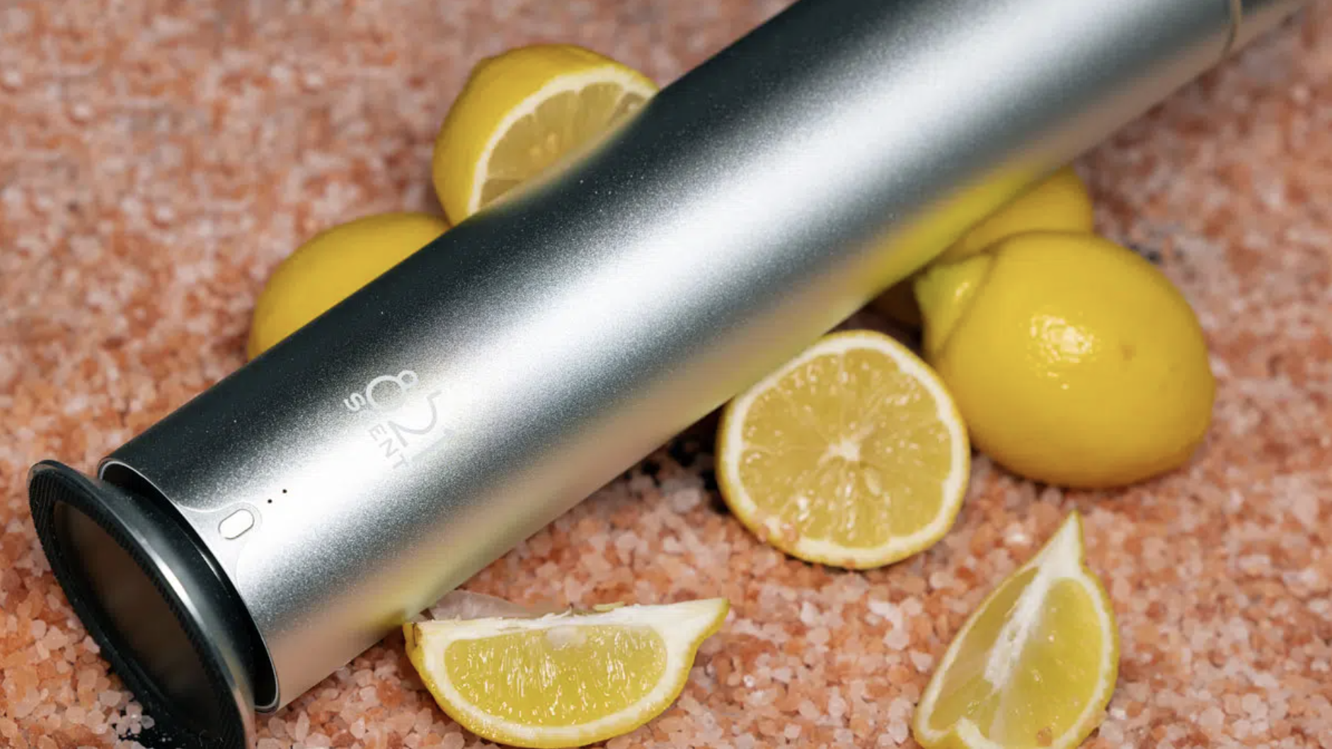 A silver portable blender lying on pink Himalayan salt with several halved and whole lemons around it.