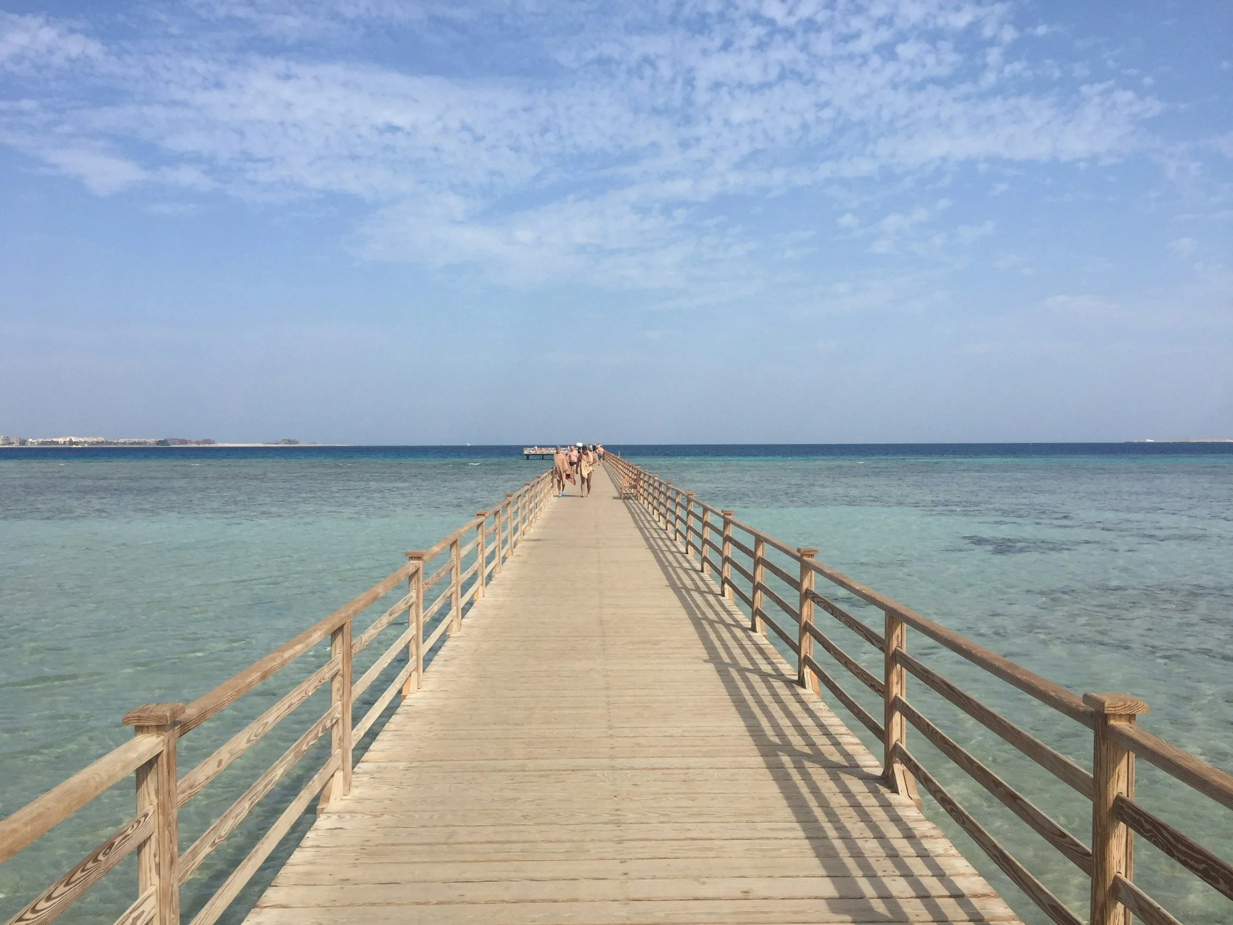 A wooden pier extends into clear, calm ocean waters with a few people walking on it, under a partly cloudy sky.