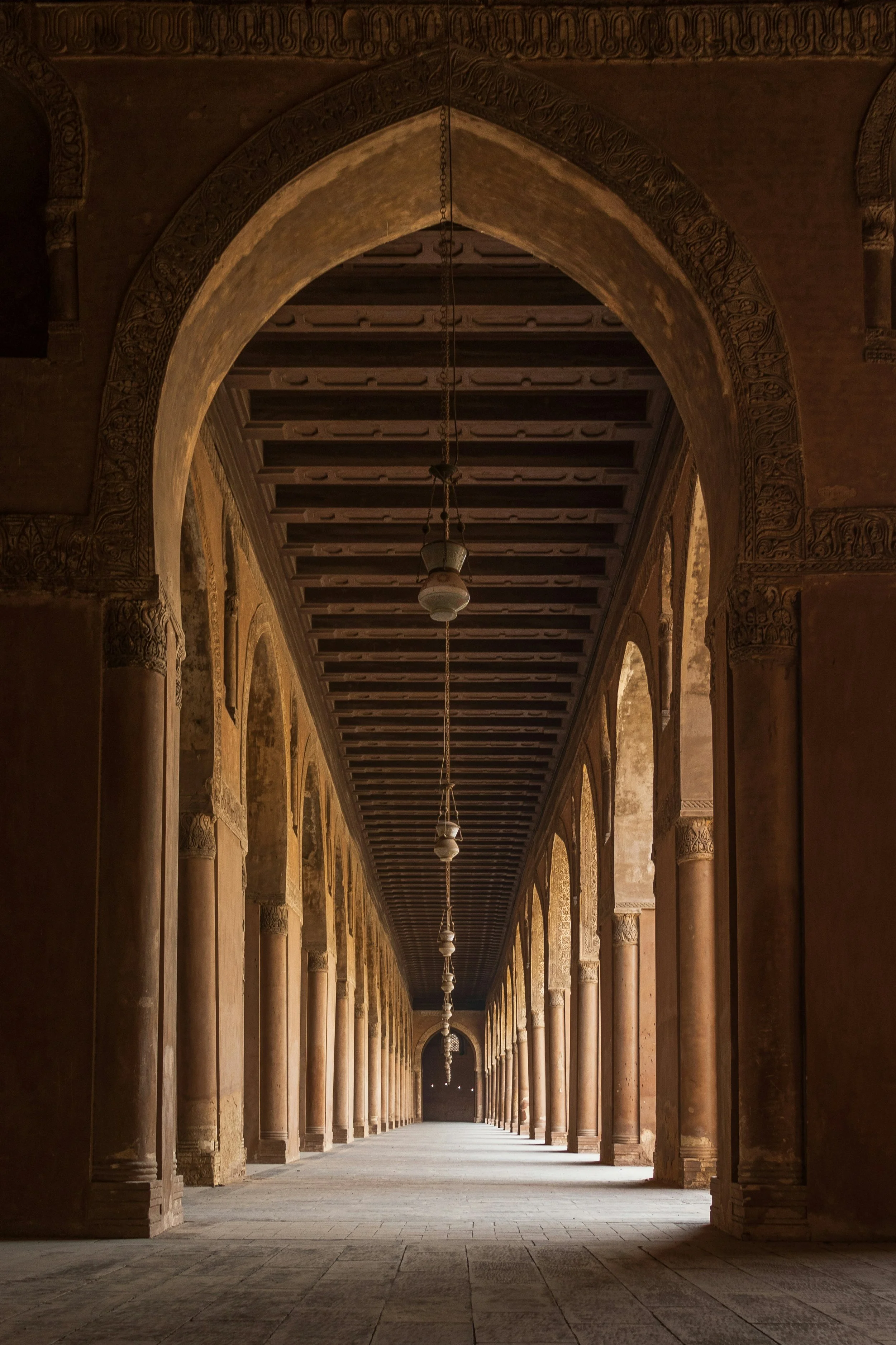 A long corridor with arches and columns, featuring intricate carvings, indoor lighting fixtures hanging from the ceiling, and a tiled floor, in what appears to be an ancient or historic building.