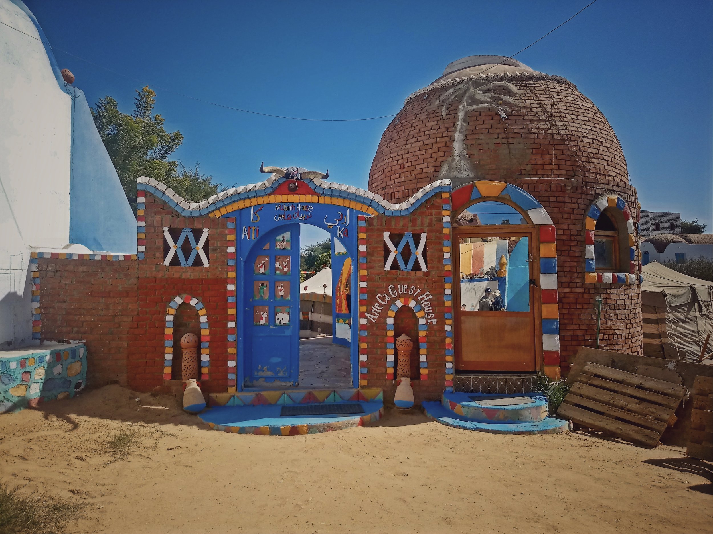 Colorful outdoor structure resembling a small house with a dome, decorated with vibrant paint and mosaic patterns, located in a sandy area with tents and trees in the background.