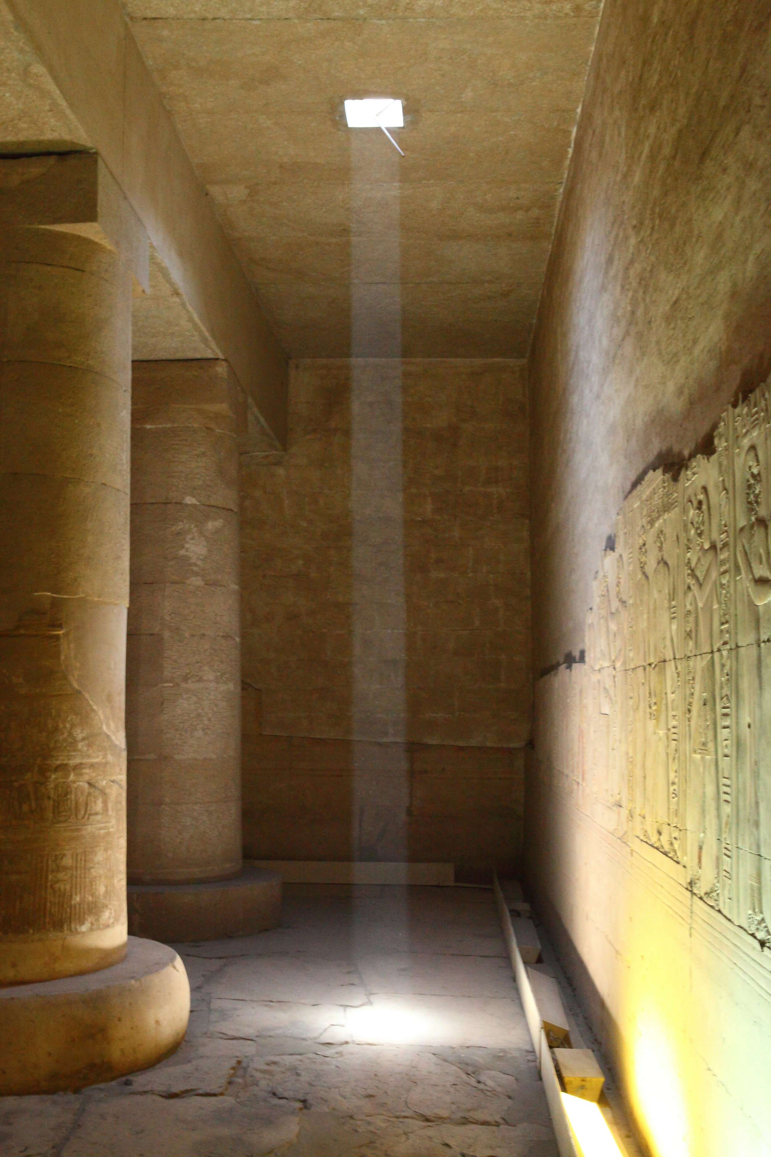 Sunlight streaming through a small opening in the ceiling of an ancient Egyptian temple, illuminating the floor and carved wall.