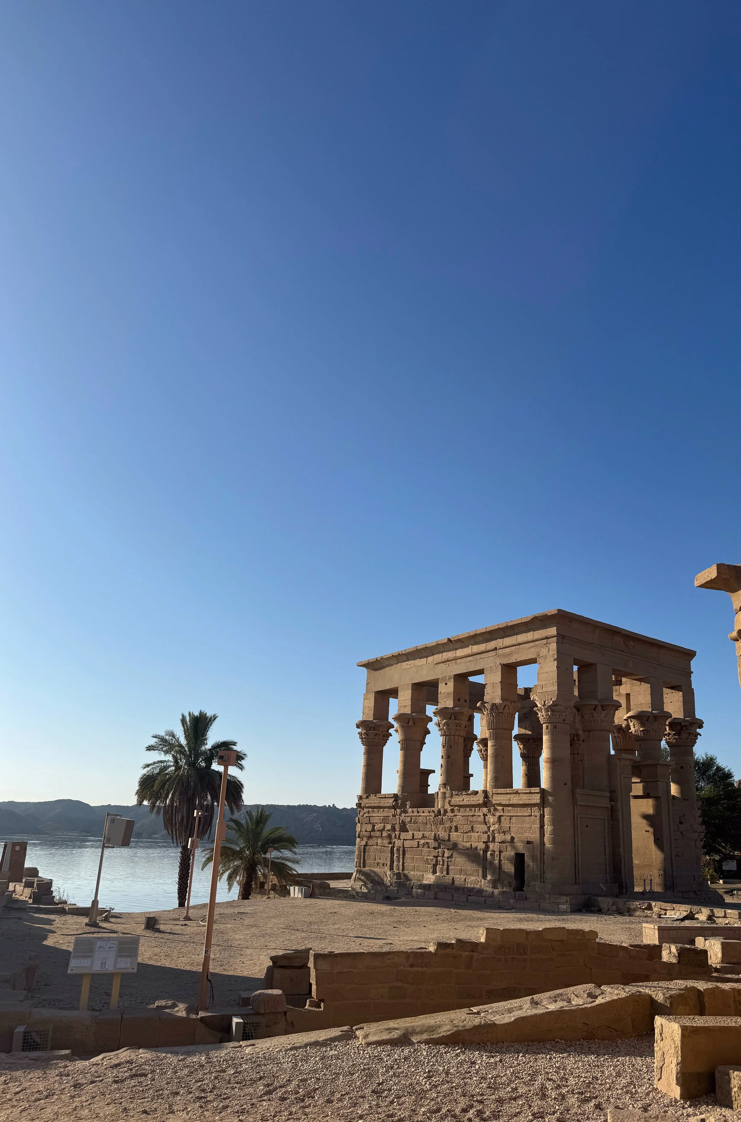 Ancient Egyptian temple ruins with a lake, palm trees, and a clear blue sky in the background.