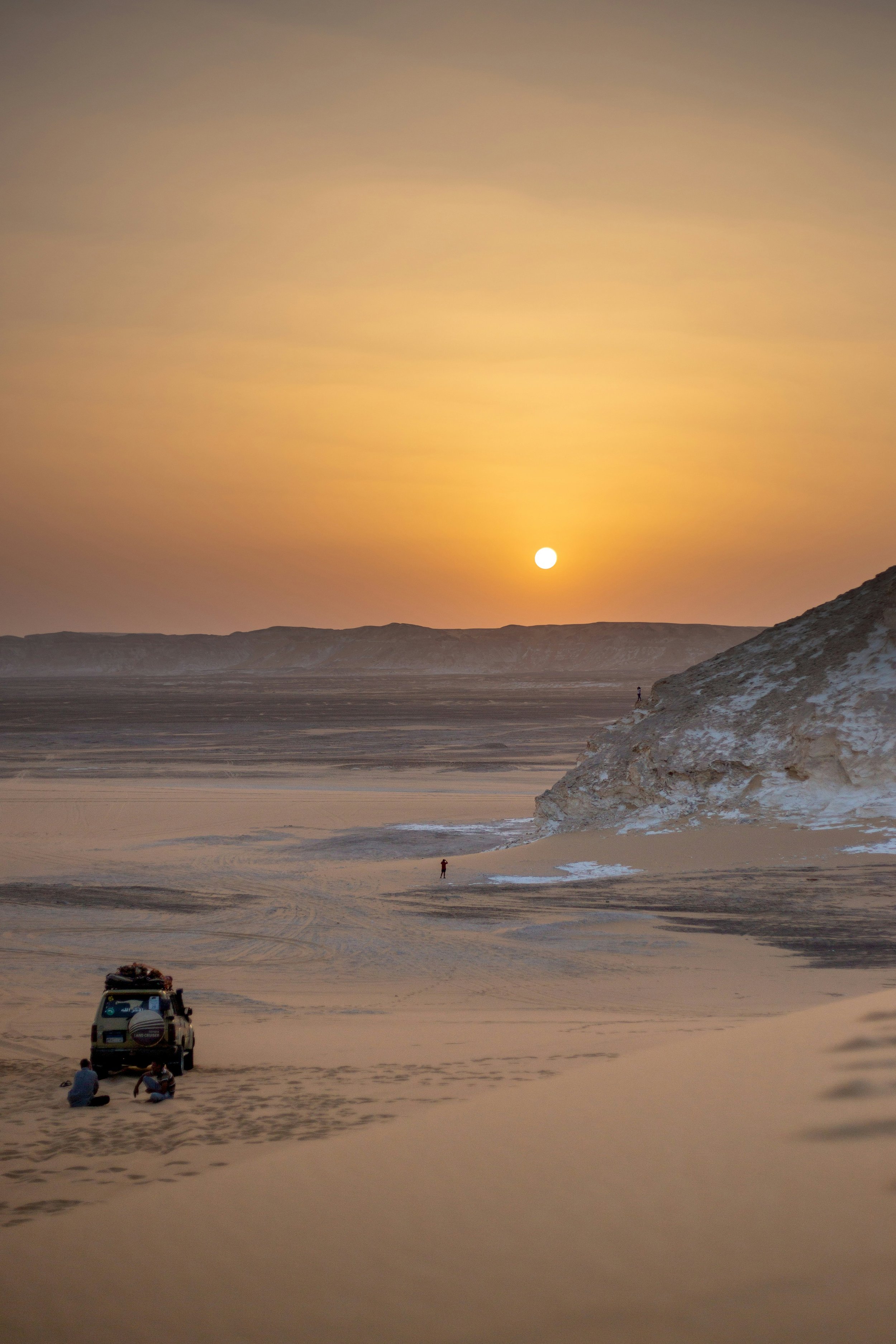 Sunset over a wide desert landscape with hills in the distance. There are a few people near a vehicle in the foreground and a person walking along the sand. The sky is clear with a soft gradient from yellow to blue.