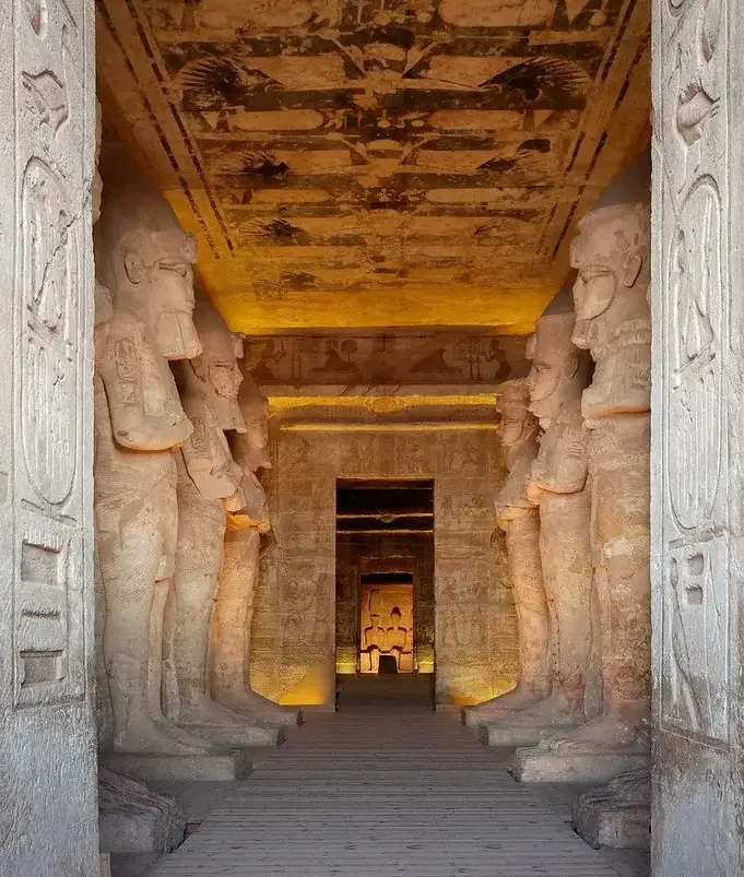 Ancient Egyptian temple corridor with large stone statues lining the walls and hieroglyphic carvings, leading to a smaller room with additional statues at the end.