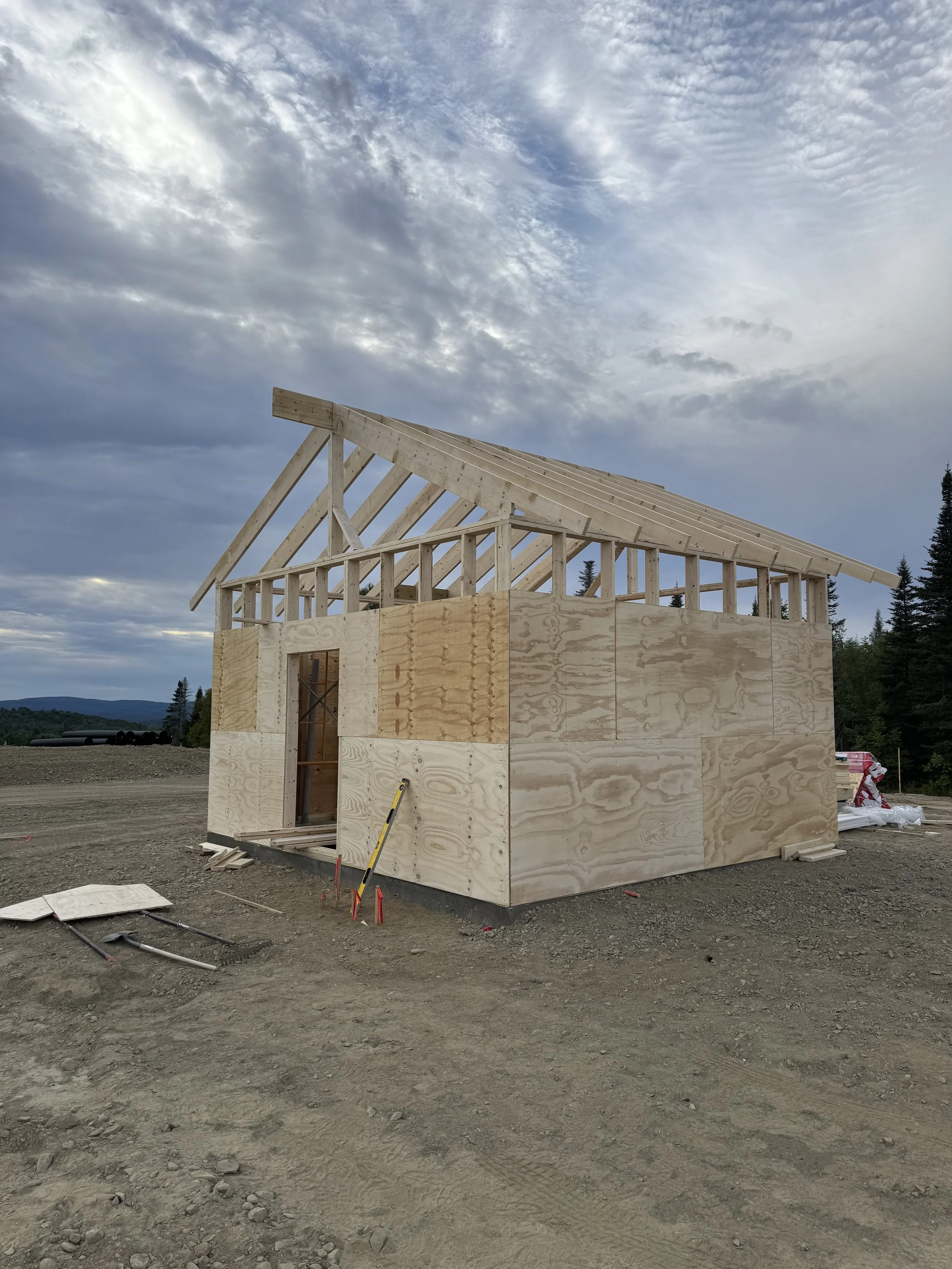 Construction site with a house frame in progress, plywood walls, and a roof structure under a cloudy sky.