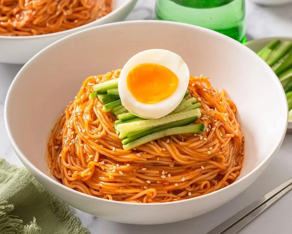 A bowl of noodles garnished with sliced cucumber, ramen egg, and sesame seeds, with additional side dishes and a drink in the background.