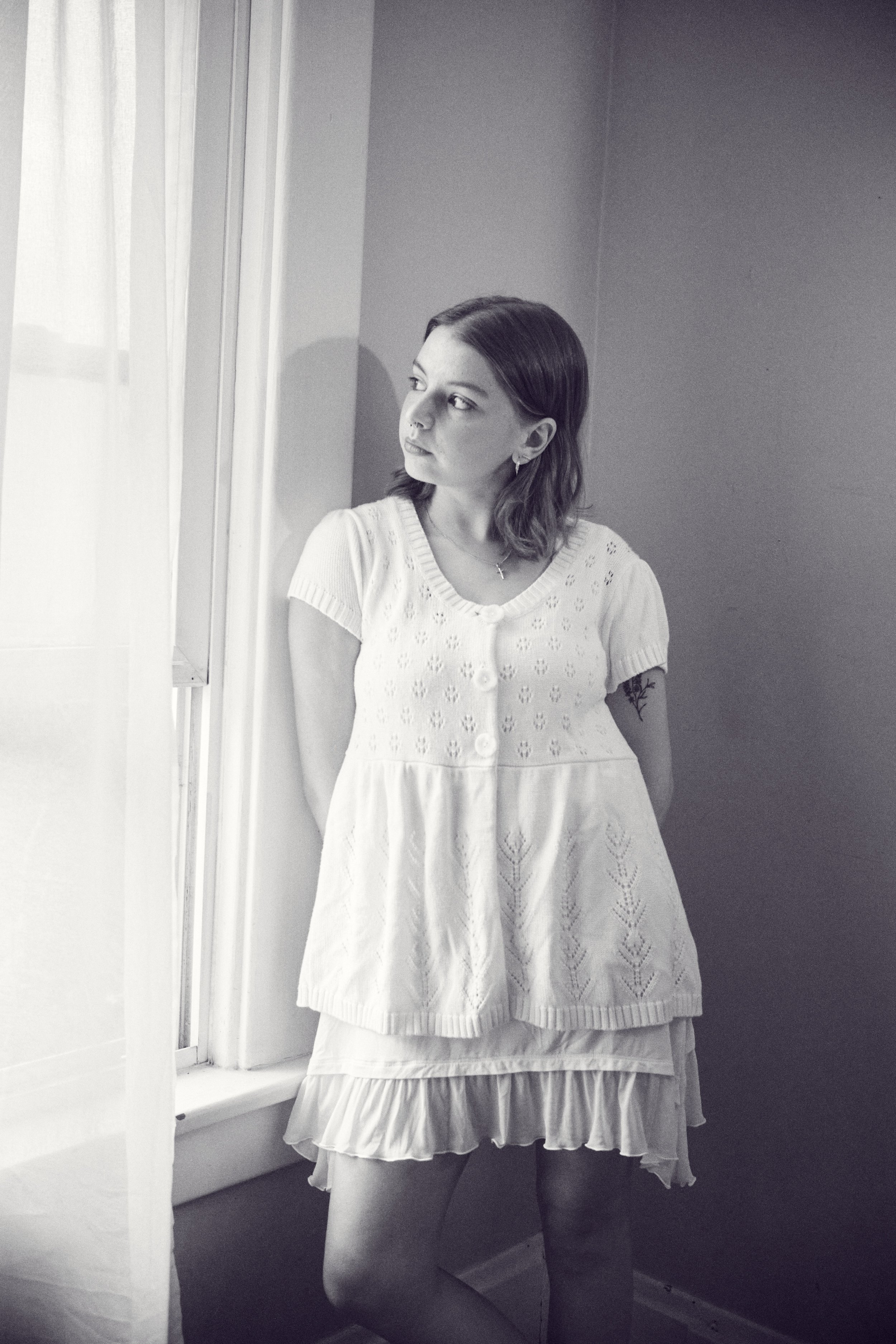 A young woman with mid-length dark hair, wearing a short-sleeved knit top and a layered skirt, standing by a window and looking outside.