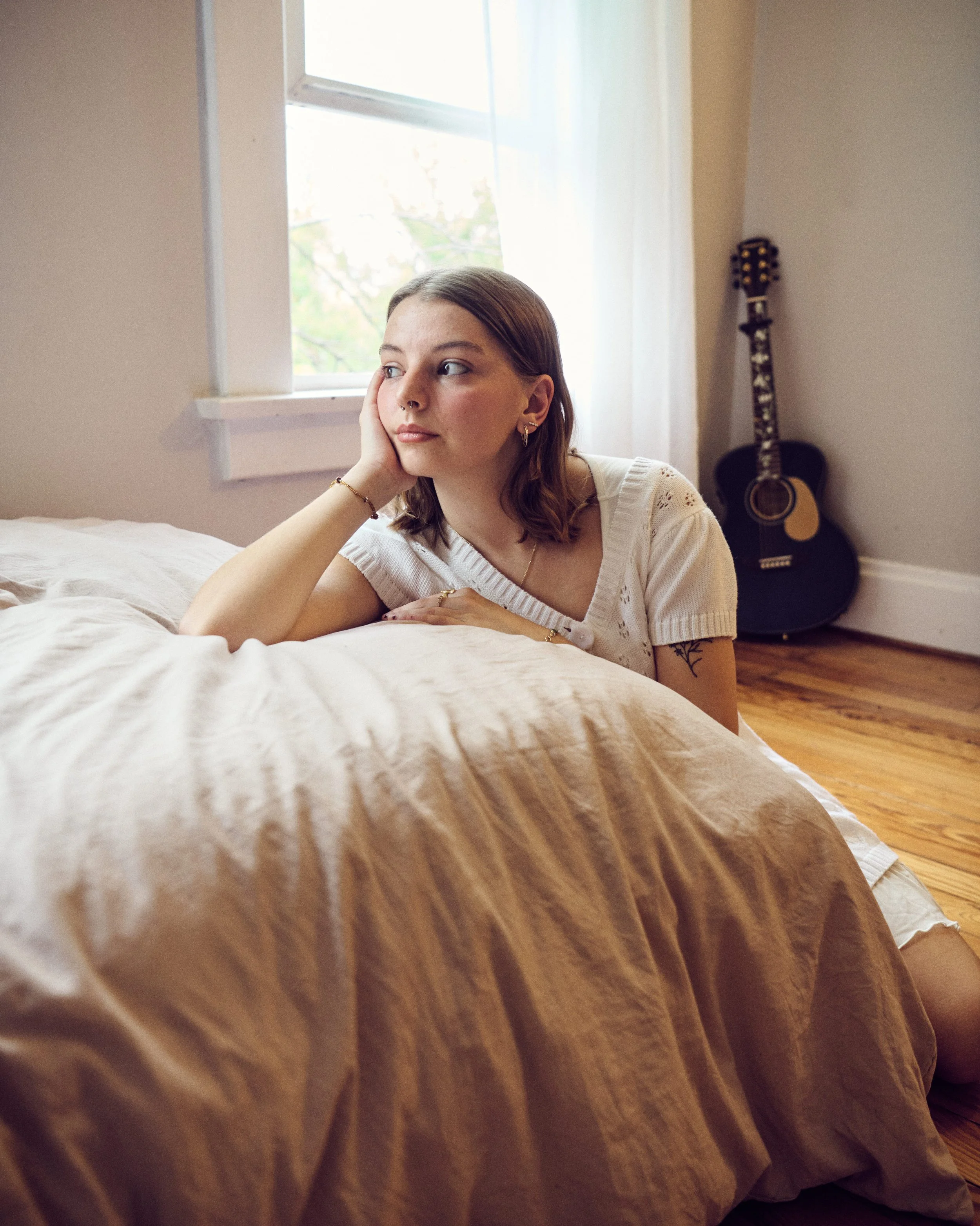A young woman with shoulder-length brown hair and jewelry is lying on her bedroom floor, resting her head on her hand, next to a bed with beige sheets. There is a window and a guitar in the background.