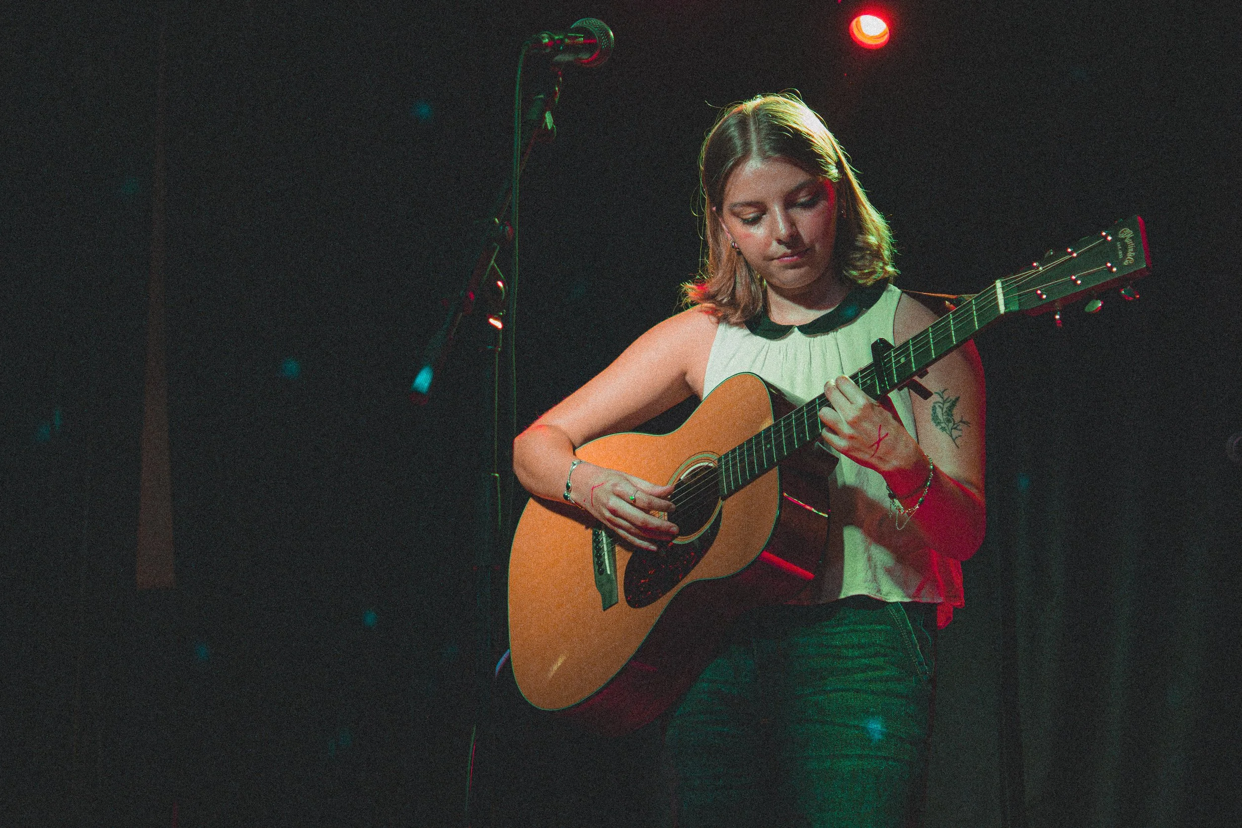 A young woman with shoulder-length hair playing an acoustic guitar on stage under red and green stage lighting.