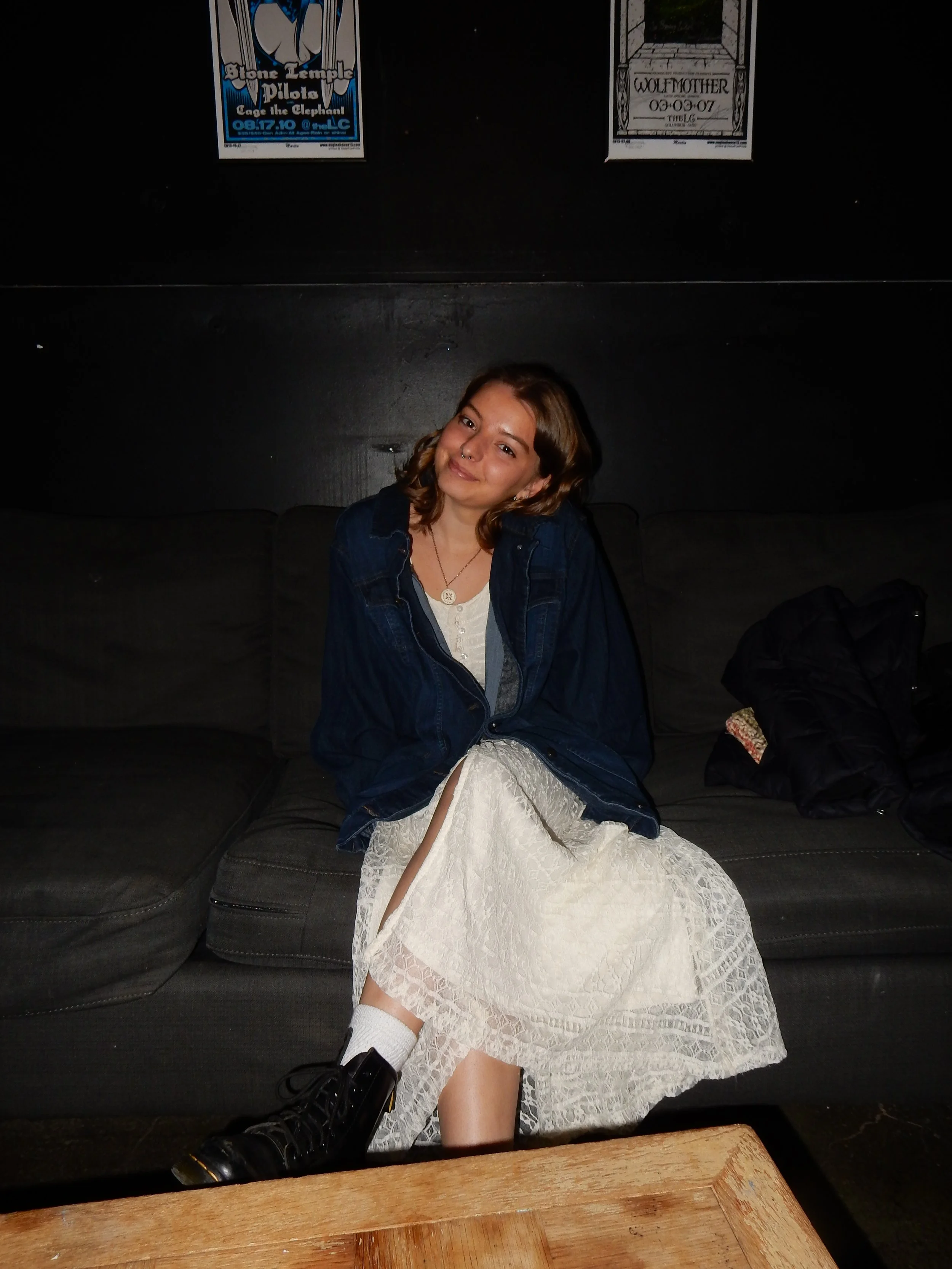 A young woman sitting on a black couch, wearing a white lace dress, a dark blue jacket, black combat boots, and white socks. She is smiling at the camera with a relaxed pose in a dimly lit room.
