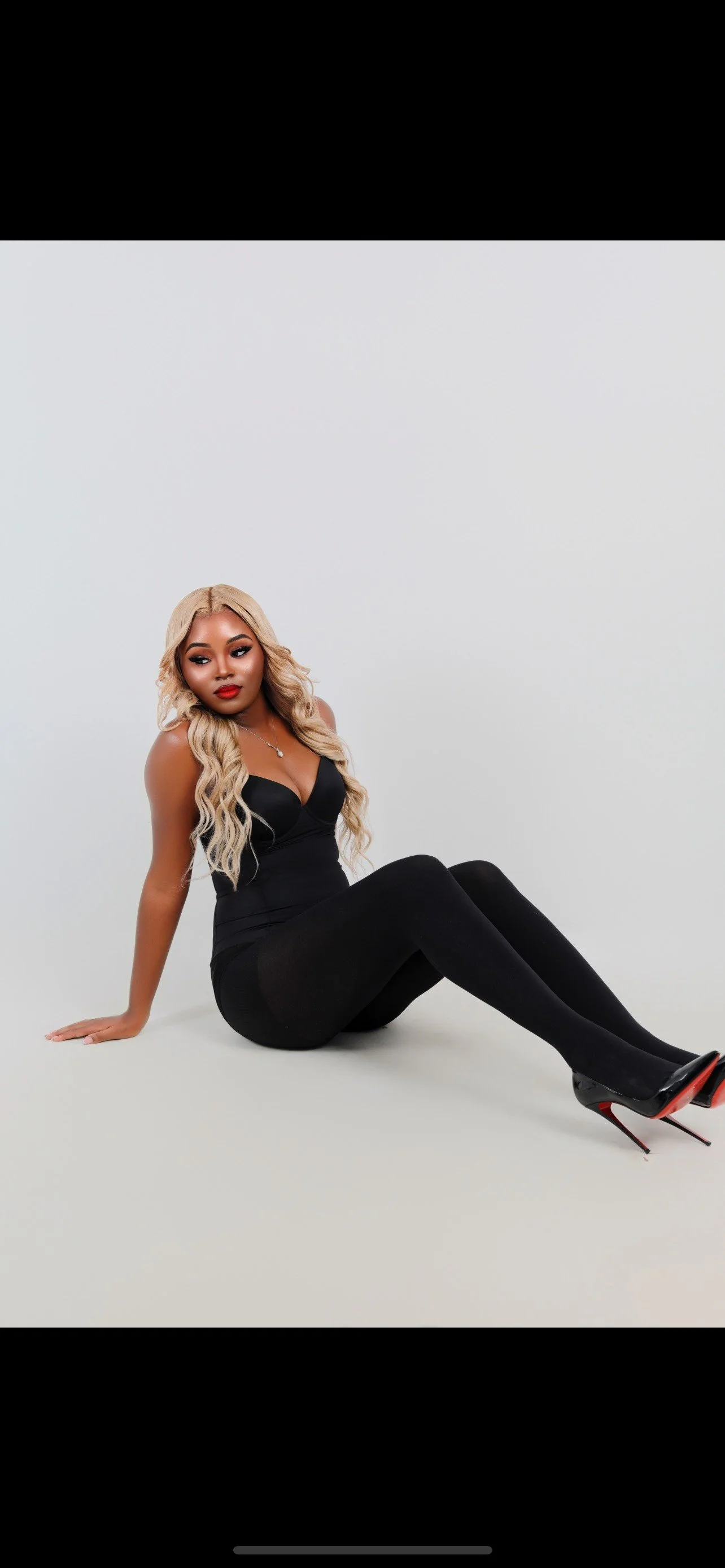 A woman with long, wavy blonde hair, wearing a black sleeveless dress, sitting on the floor with her legs extended and crossed at the ankles. She has red lipstick and makeup, and is wearing black high heels.