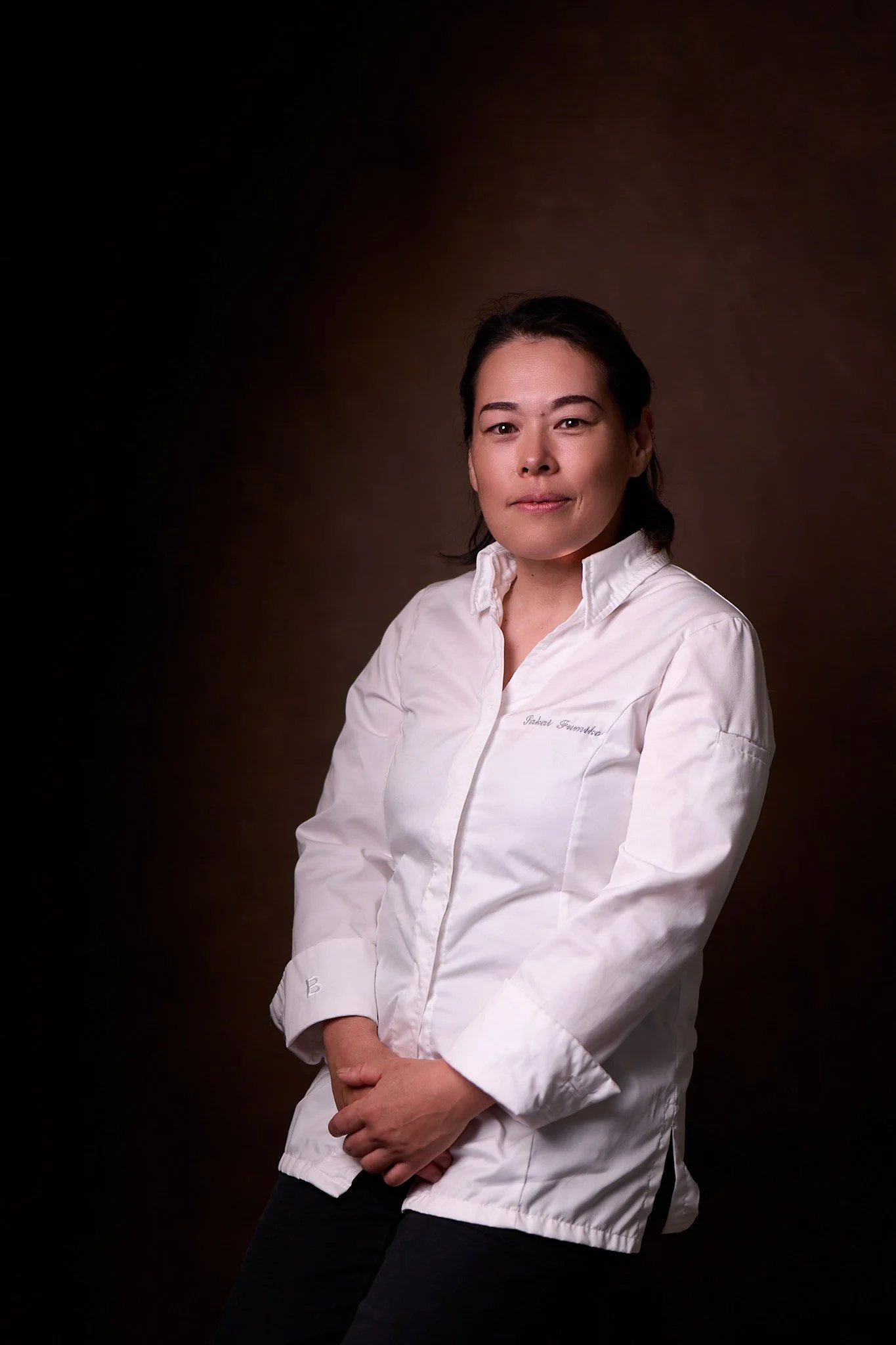 Una donna con capelli scuri riccioli indossa una giacca bianca e si trova di fronte a uno sfondo sfumato marrone scuro.