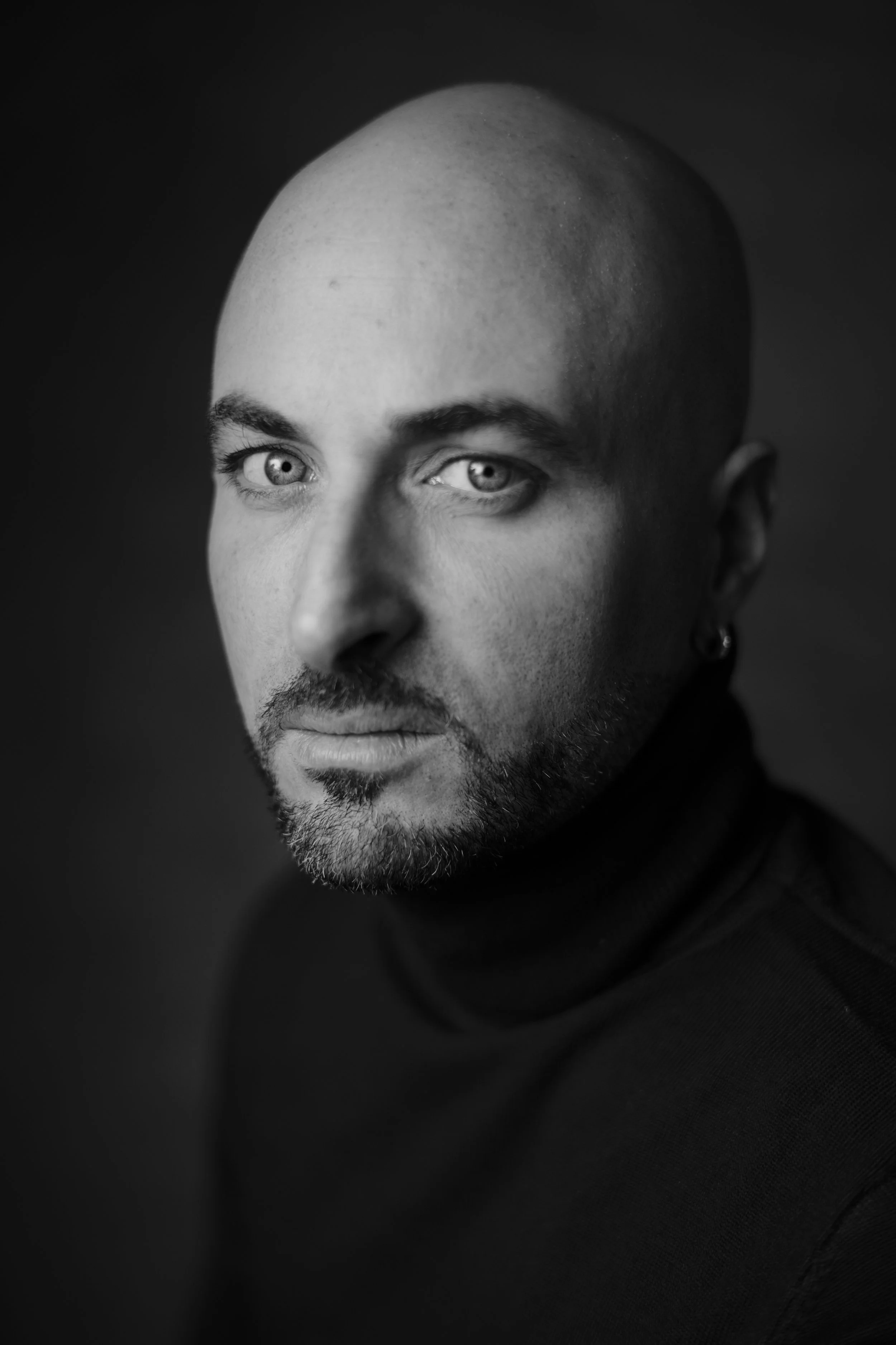 Ritratto in bianco e nero di un uomo con testa rasata, occhi chiari, barba e orecchino, indossa un maglione a collo alto neri.