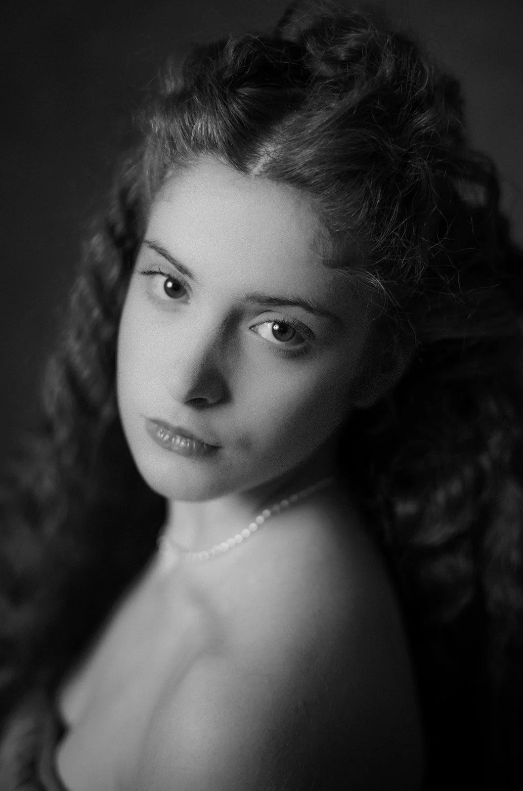 Ritratto di una giovane donna con capelli ricci scuri, ma in bianco e nero, che guarda verso l'obiettivo, indossa una collana e ha un'espressione serena.