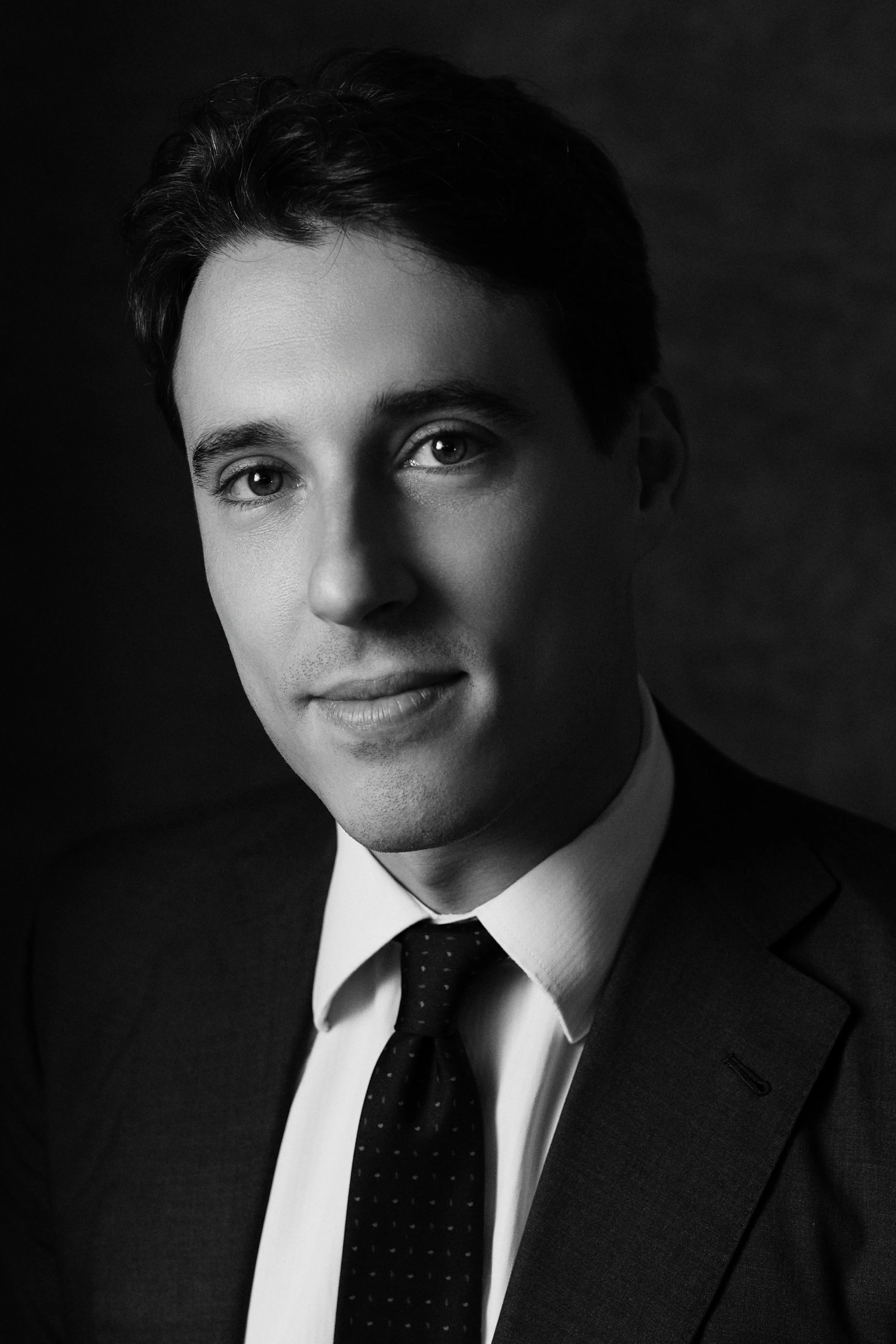 Ritratto in bianco e nero di un uomo con capelli scuri, vestito con giacca e cravatta, con un'espressione seria e sorriso leggero.