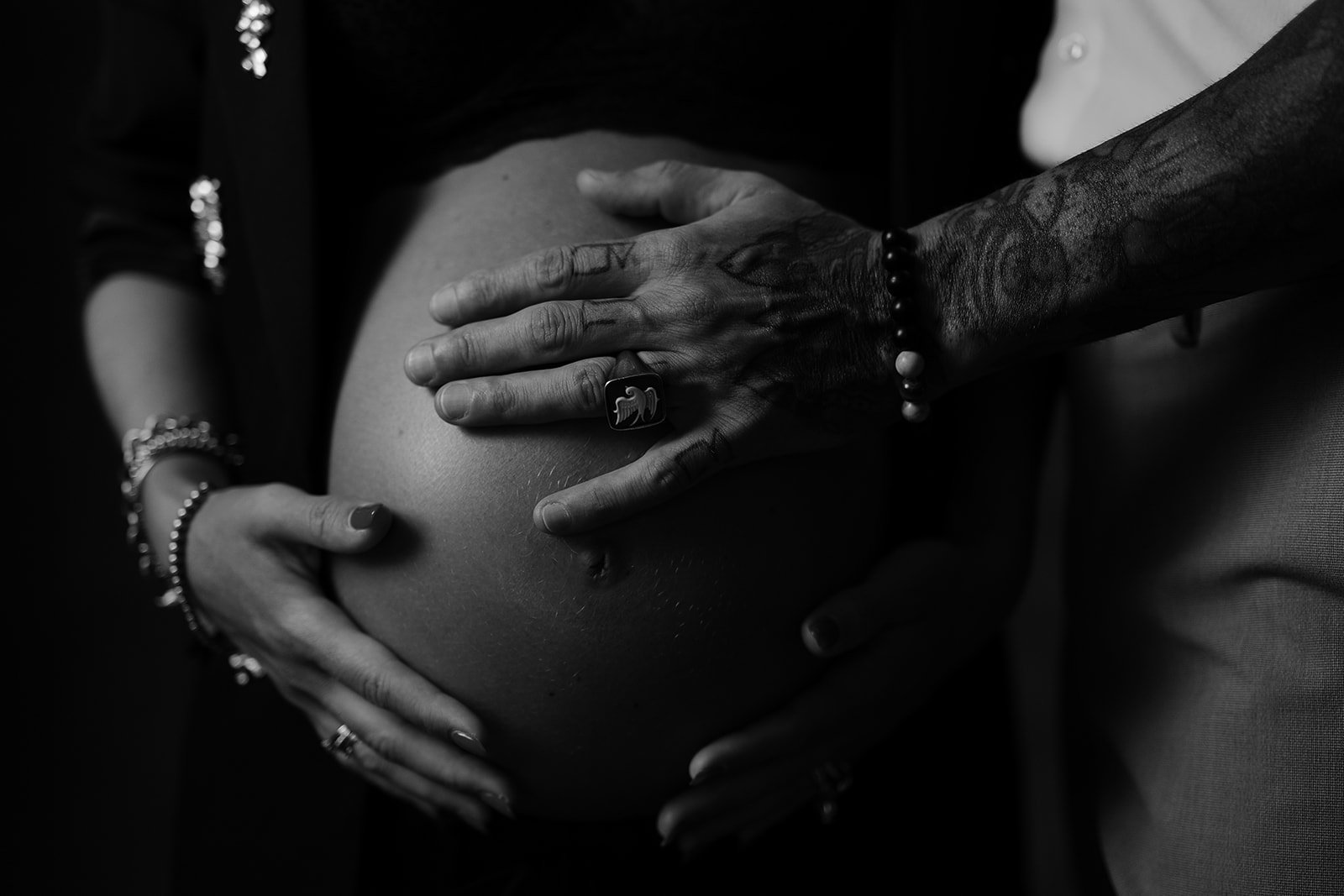Due mani tatuate e adornate con braccialetti e anelli, custodiscono e sostengono un pancione di una donna incinta in primo piano, in una scena in bianco e nero.