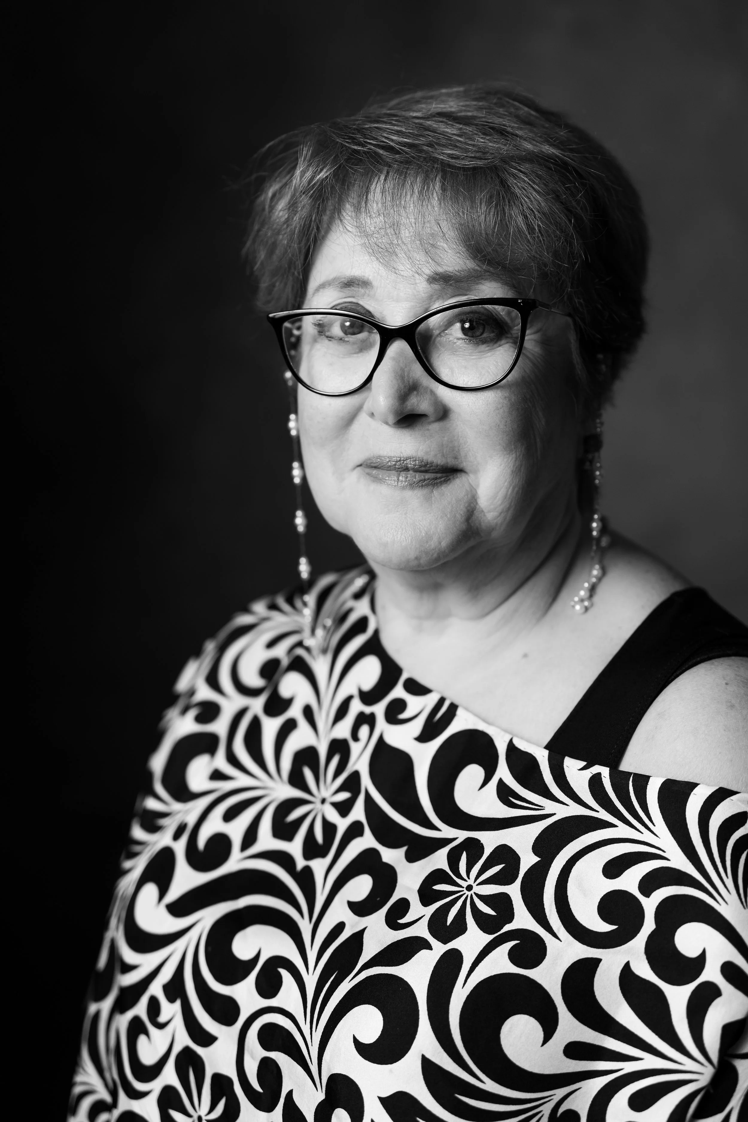 Ritratto in bianco e nero di una donna anziana con occhiali neri, orecchini pendenti e vestito con motivo floreale.