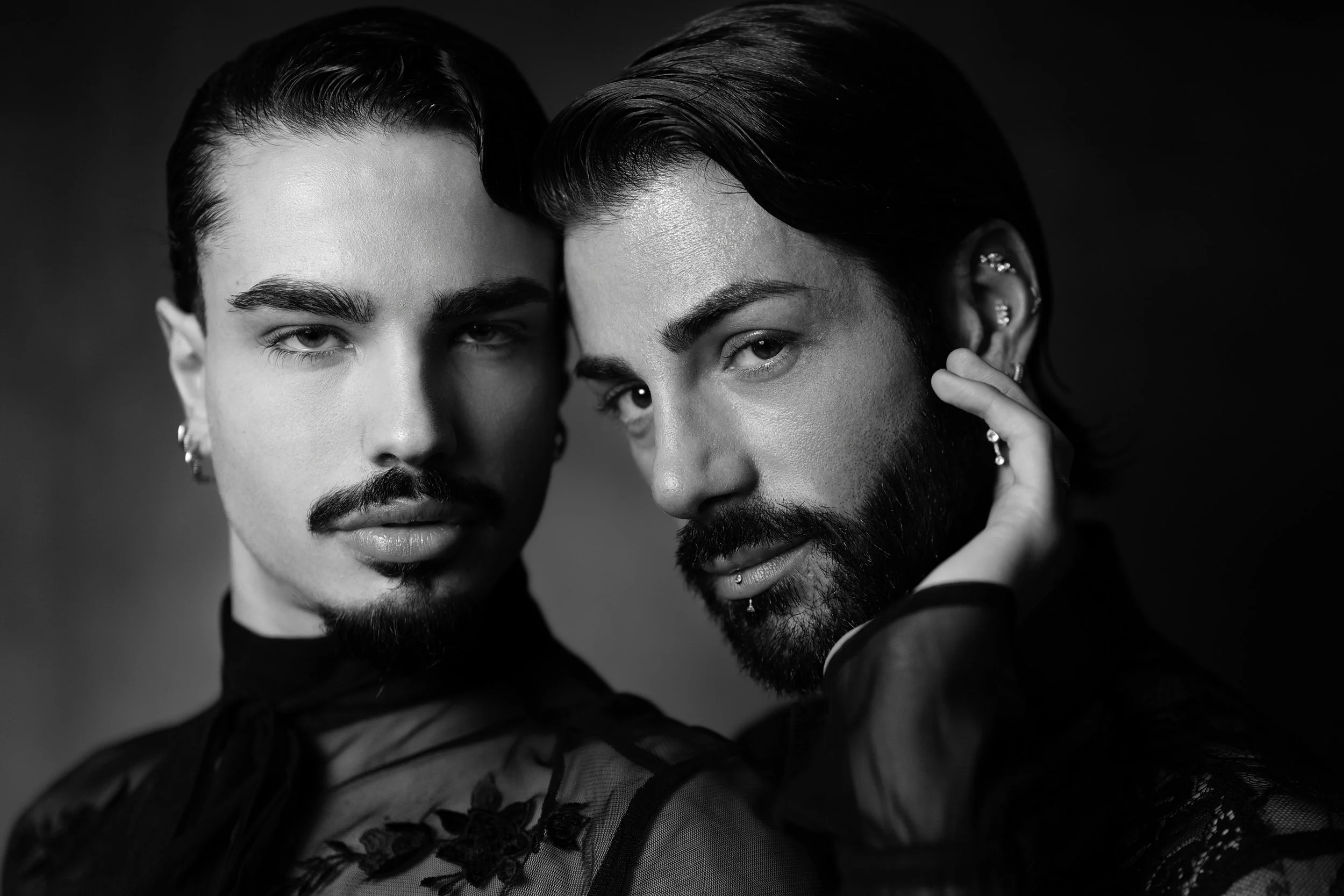 Due uomini con tratti androgini, uno con barba e l'altro senza, si guardano intensamente in un ritratto in bianco e nero.
