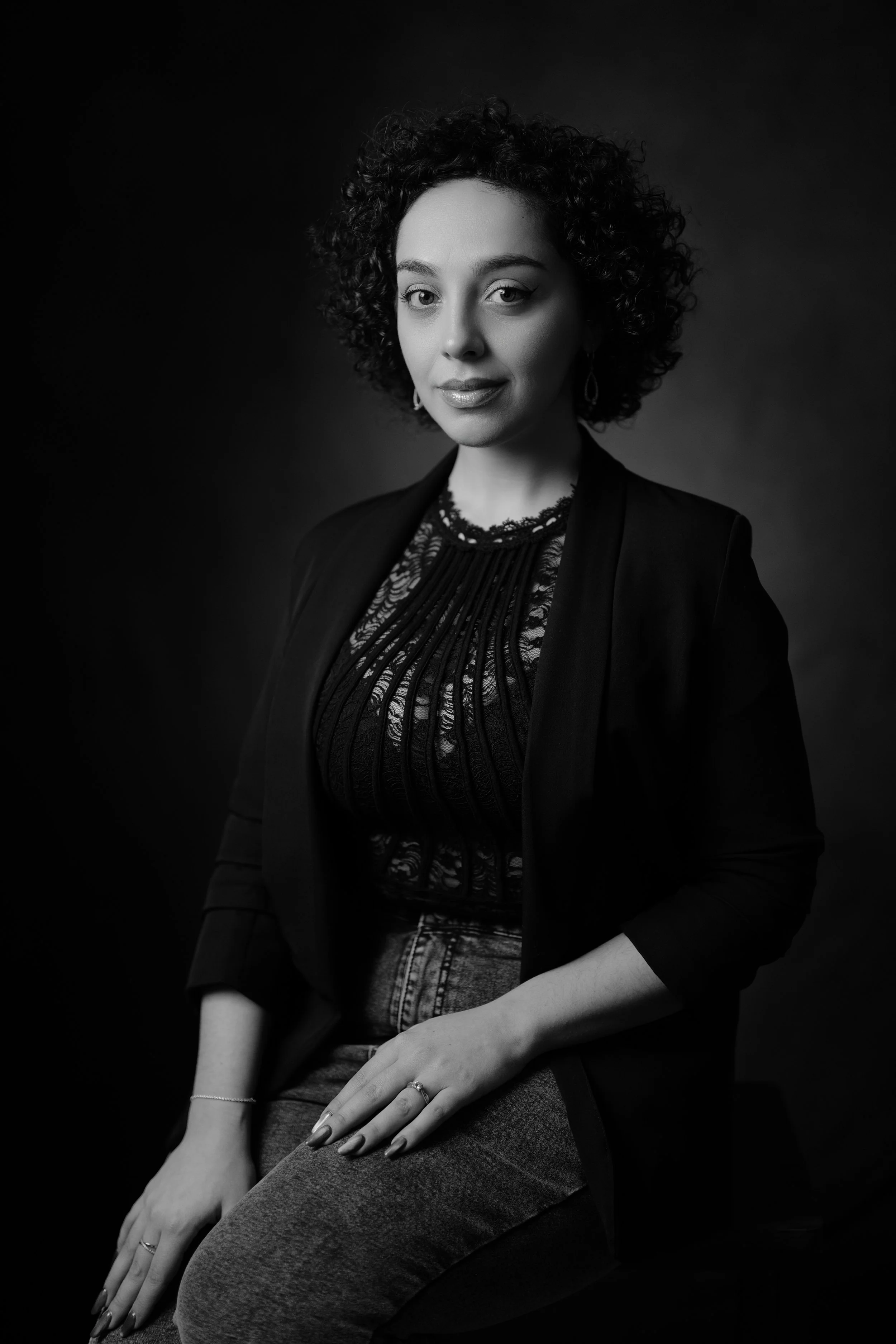 Ragazza con capelli riccioli scuri, indossa una giacca nera e una blusa di pizzo, seduta su una sedia, foto in bianco e nero.
