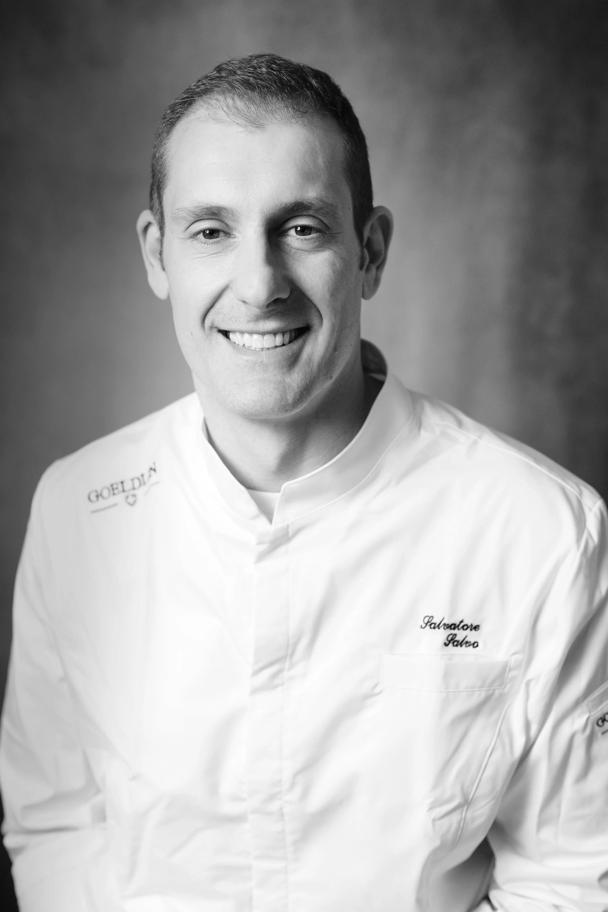 Foto in bianco e nero di uno chef sorridente con giacca bianca, scritto 'Salvatore Galato', contro uno sfondo sfocato.