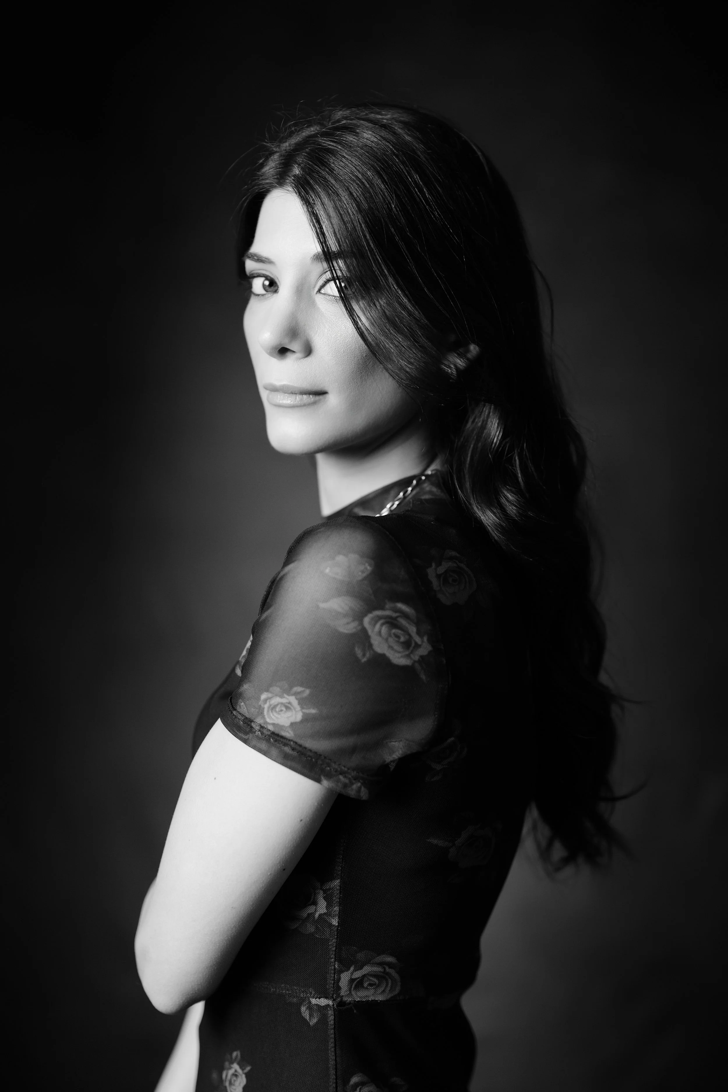 Ritratto in bianco e nero di una donna con capelli lunghi e scuri, vestita con un top nero con maniche di tessuto trasparente decorato con rose, con un aspetto elegante e sereno.