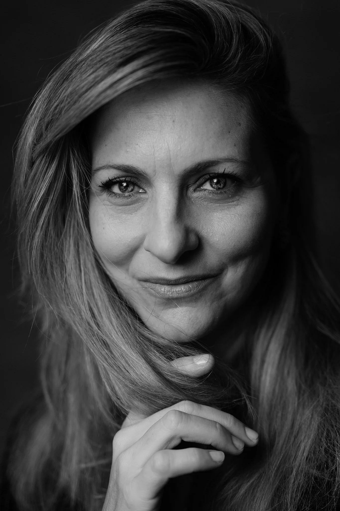 Ritratto in bianco e nero di una donna con capelli lunghi e occhi chiari, che sorride leggermente con la mano sotto il mento.