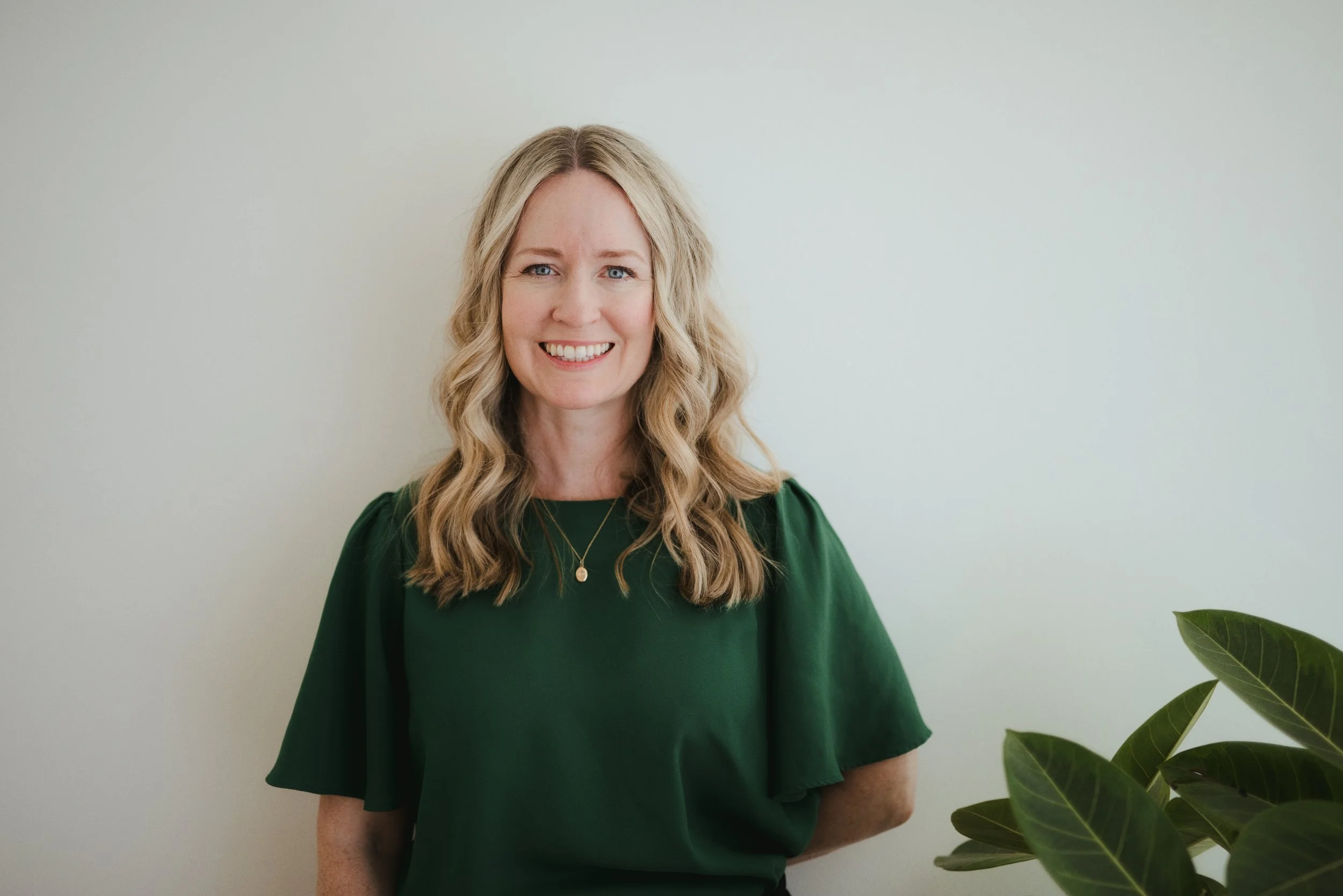 Woman in green shirt looking at camera and smiling with one arm behind back resting on wall
