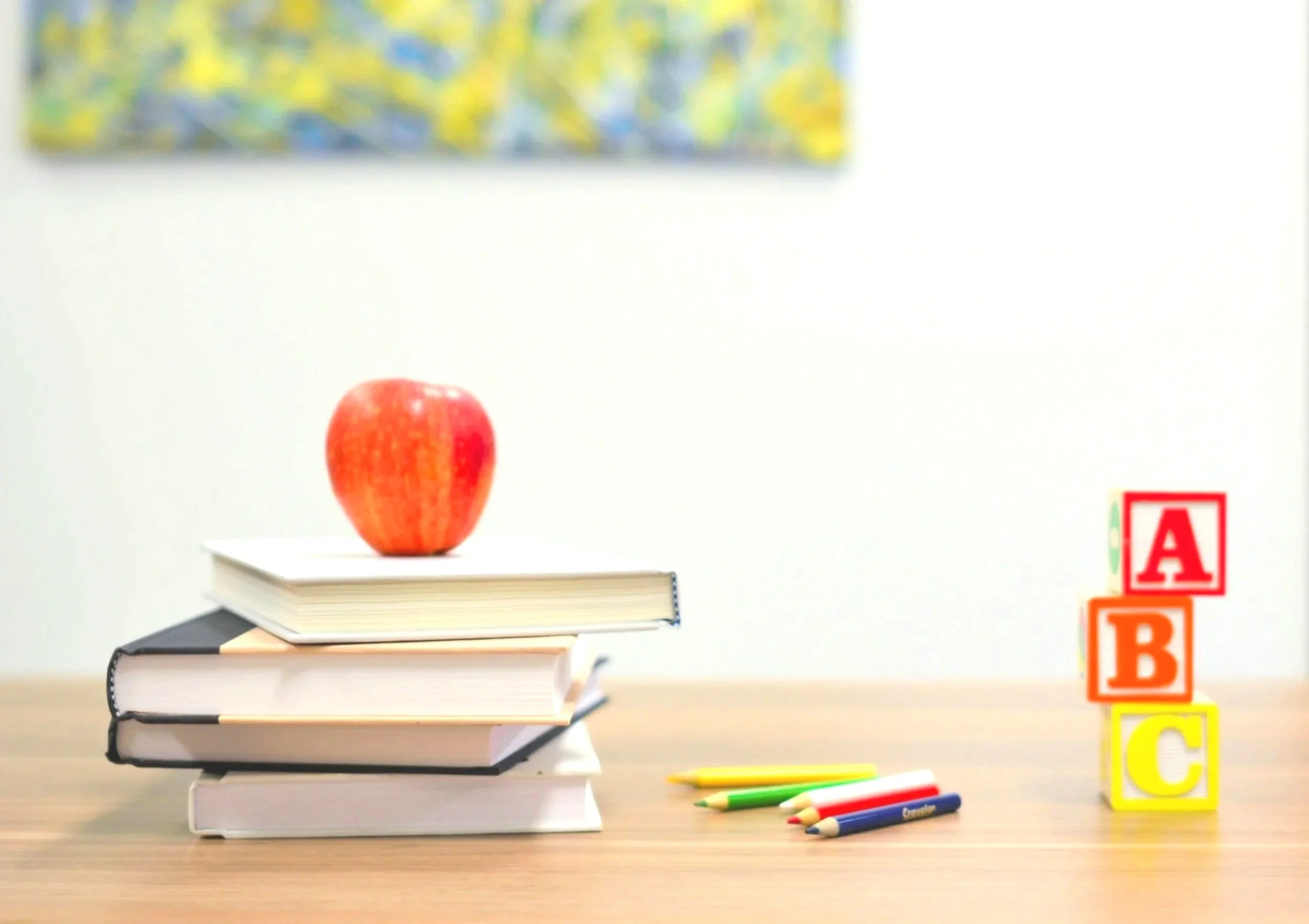 A red apple on top of three stacked books on a wooden table with yellow, green, red, and blue pens. To the right, a stack of colorful blocks spelling out A, B, and C.