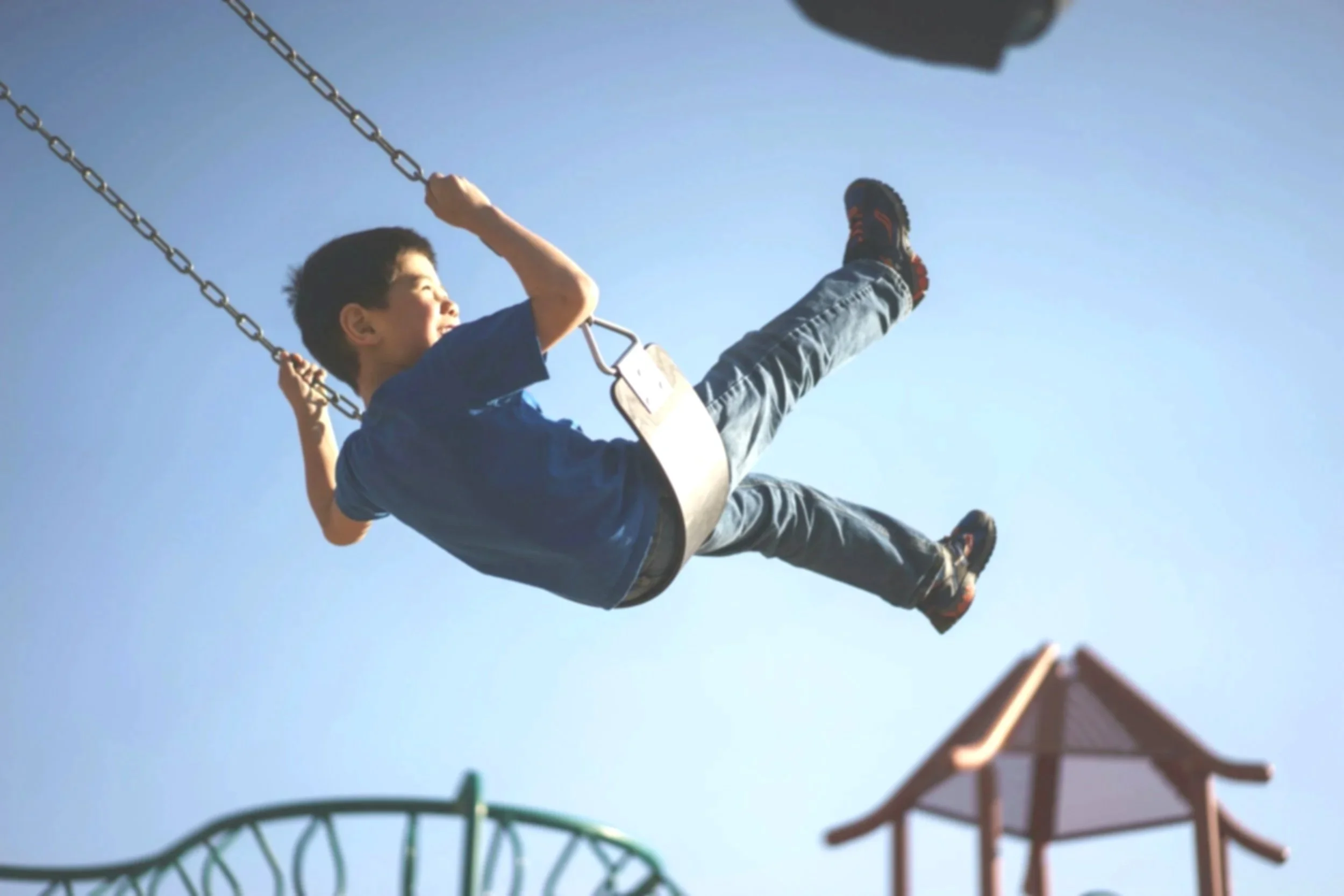 Child boy swinging on a playground swing set against a clear blue sky.
