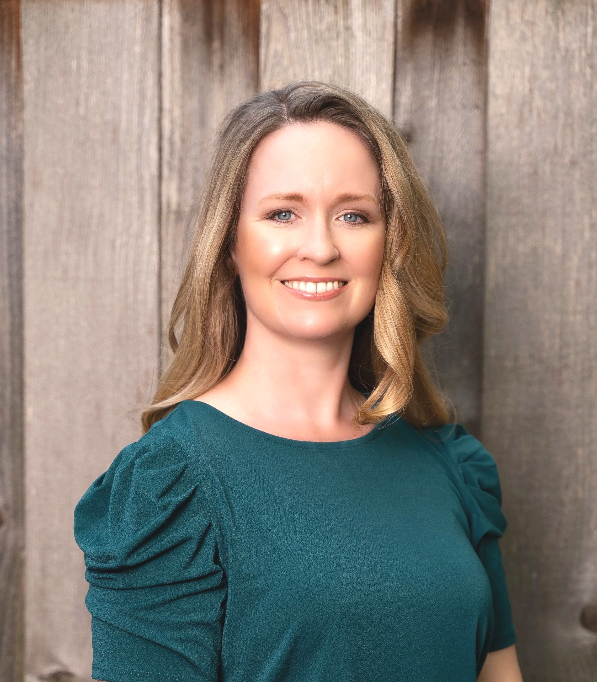 A woman with blonde hair wearing a teal top, smiling, standing against a wooden background.