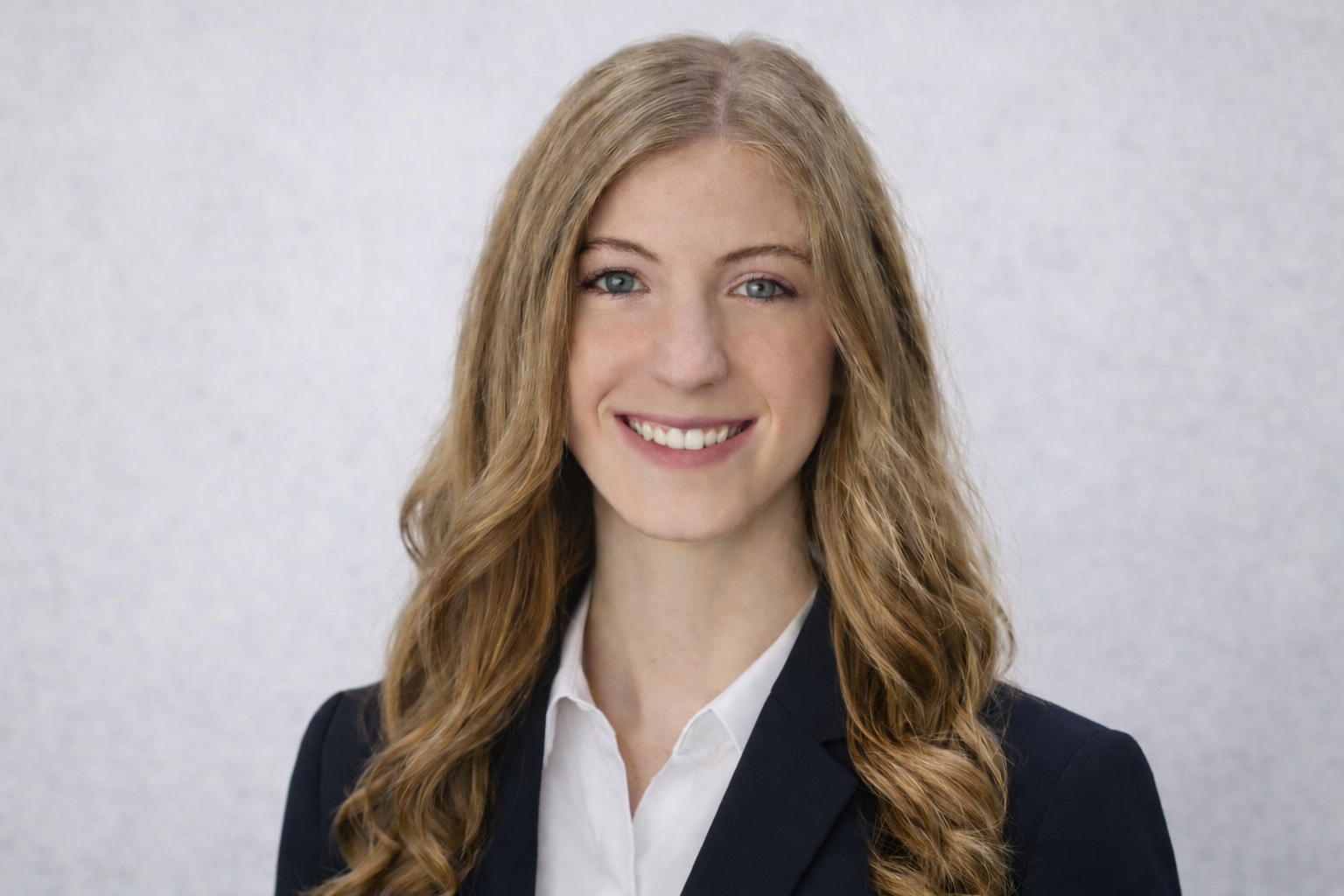 Professional woman with long curly red hair, wearing a white shirt and a dark blazer, smiling against a plain light gray background.