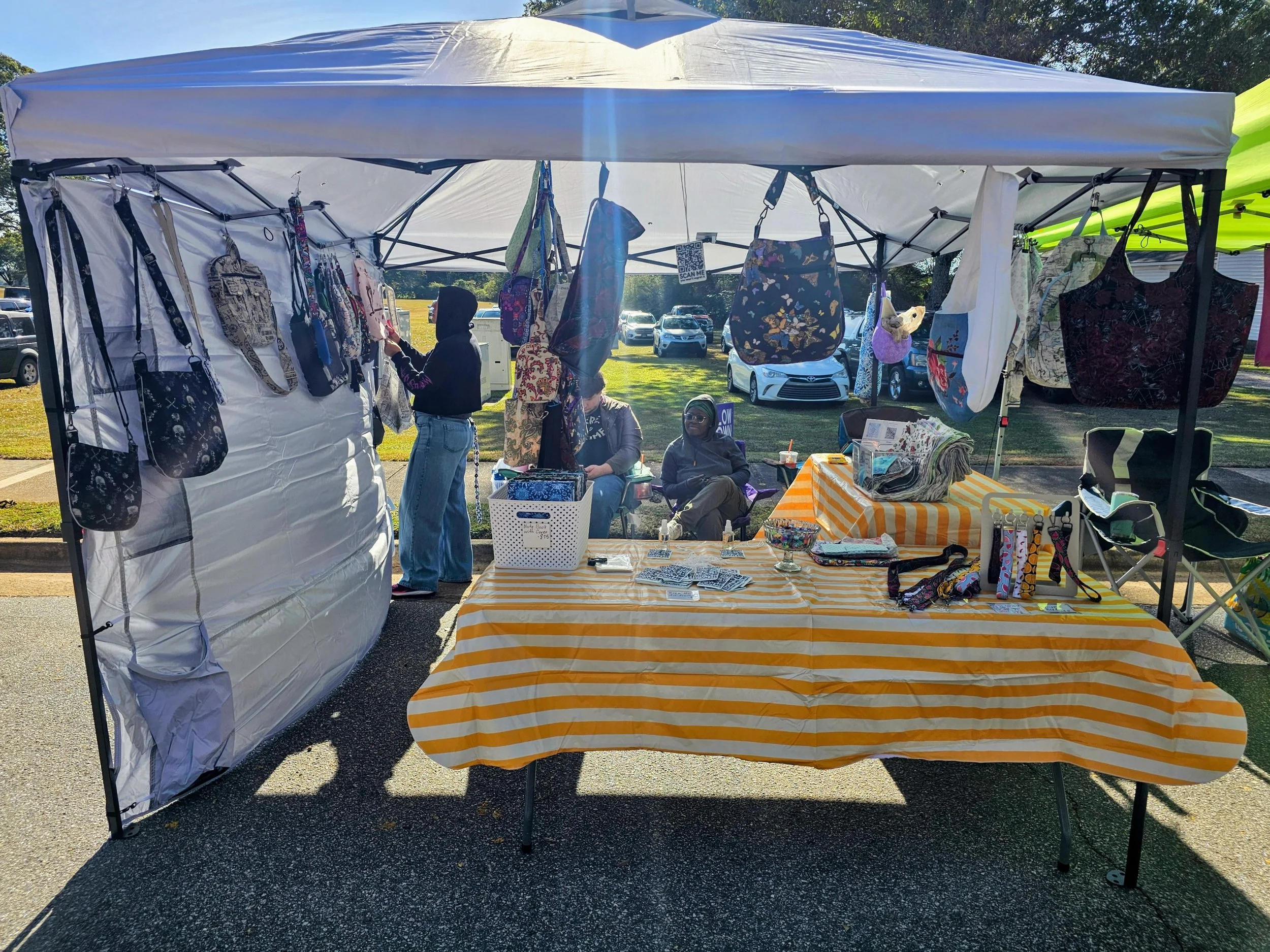 A market stall at an outdoor fair with bags, wallets, and fabric items displayed beneath a white canopy tent. Two women are sitting and chatting behind the table, while another person is browsing. Cars are parked in the background on a sunny day.