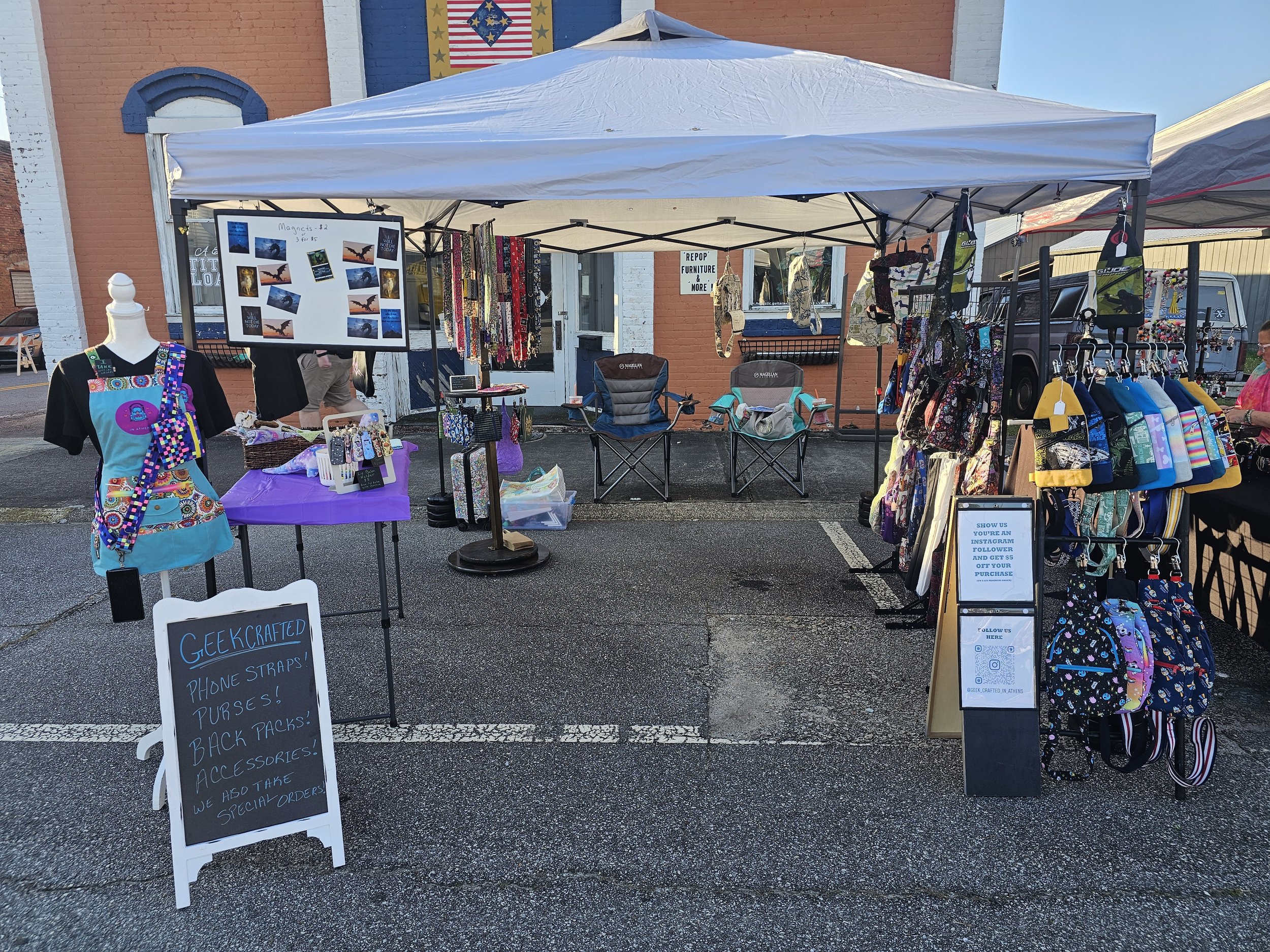 An outdoor market stall with a white canopy. The stall displays various accessories including bags, phone straps, and keychains. There are folding chairs and a purple tablecloth with items for sale. Signs indicate the items sold, including geek-crafted straps, phone cases, purses, back packs, and accessories.