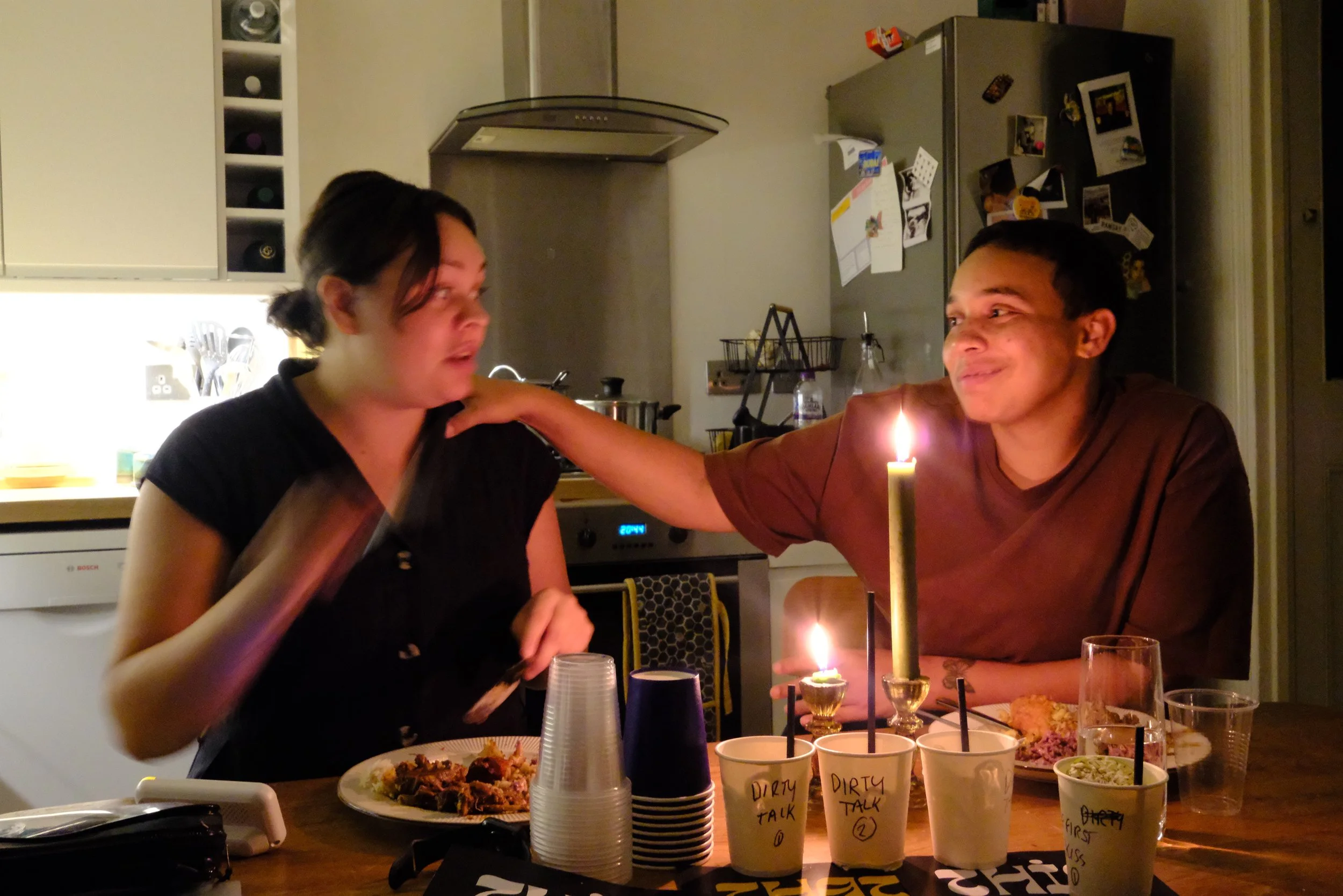 Two people sitting at a dinner table with candles, food, and drinks, engaging in conversation, in a cozy indoor setting.