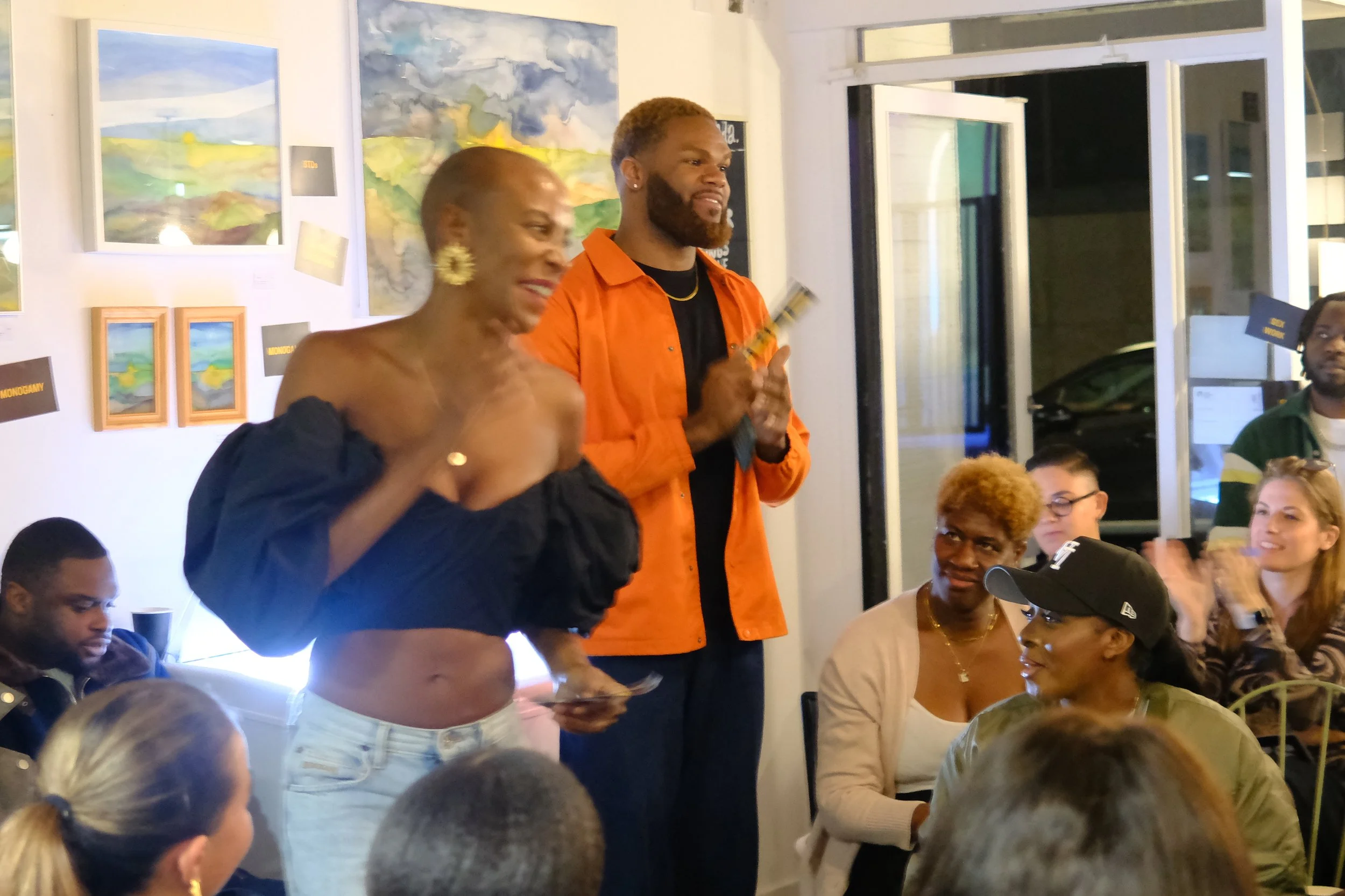 A group of people gathered indoors, with two individuals standing and speaking to the audience. The woman on the left is wearing an off-the-shoulder black top and white jeans, smiling and gesturing. The man on the right is wearing an orange shirt over a black shirt, holding what appears to be a microphone or musical instrument. The audience is seated, clapping, and attentively listening, with artwork hanging on the wall behind them.