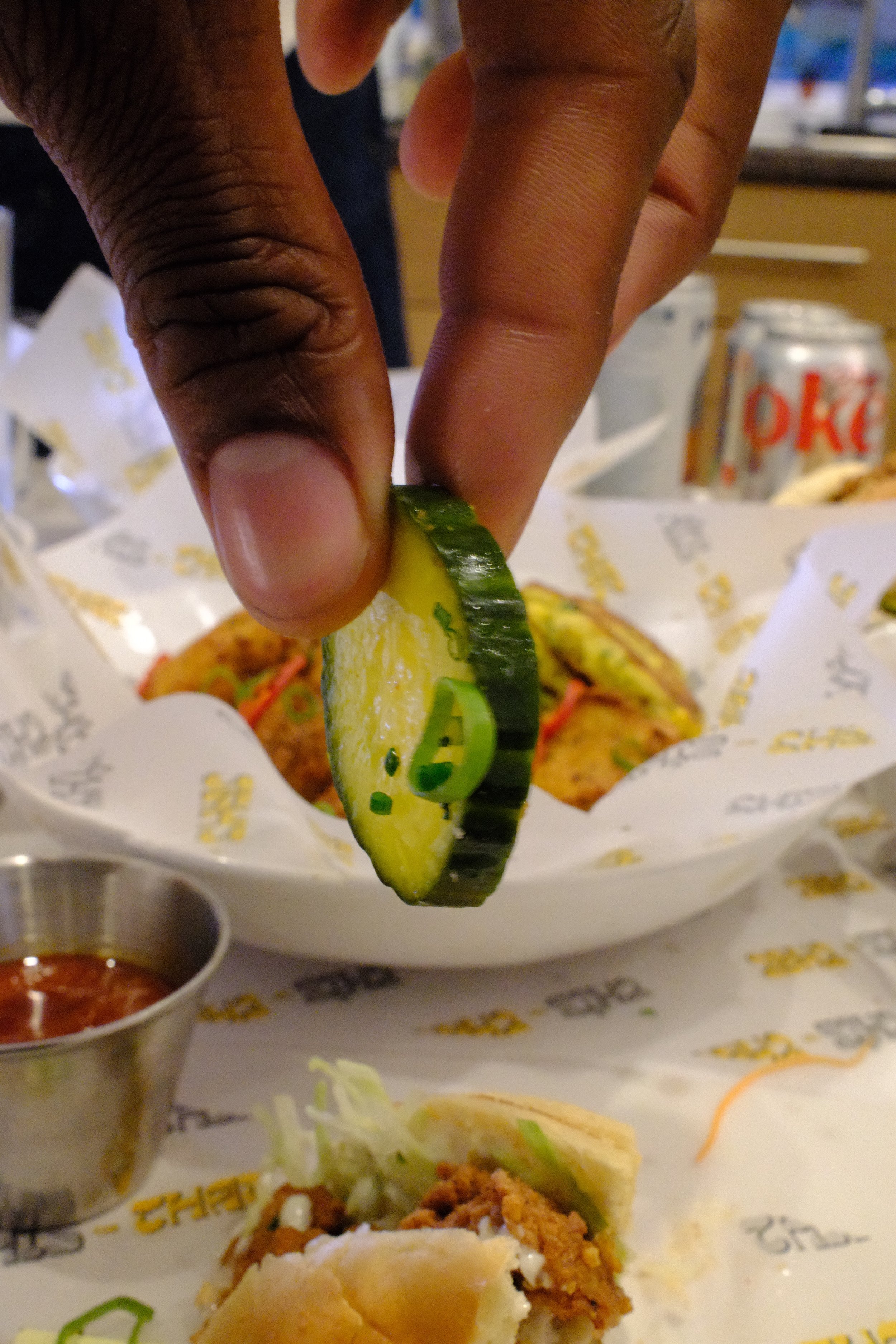 Close-up of a hand placing a slice of cucumber on a plate of fried chicken, sandwich bread, and dipping sauces on a table at a restaurant.
