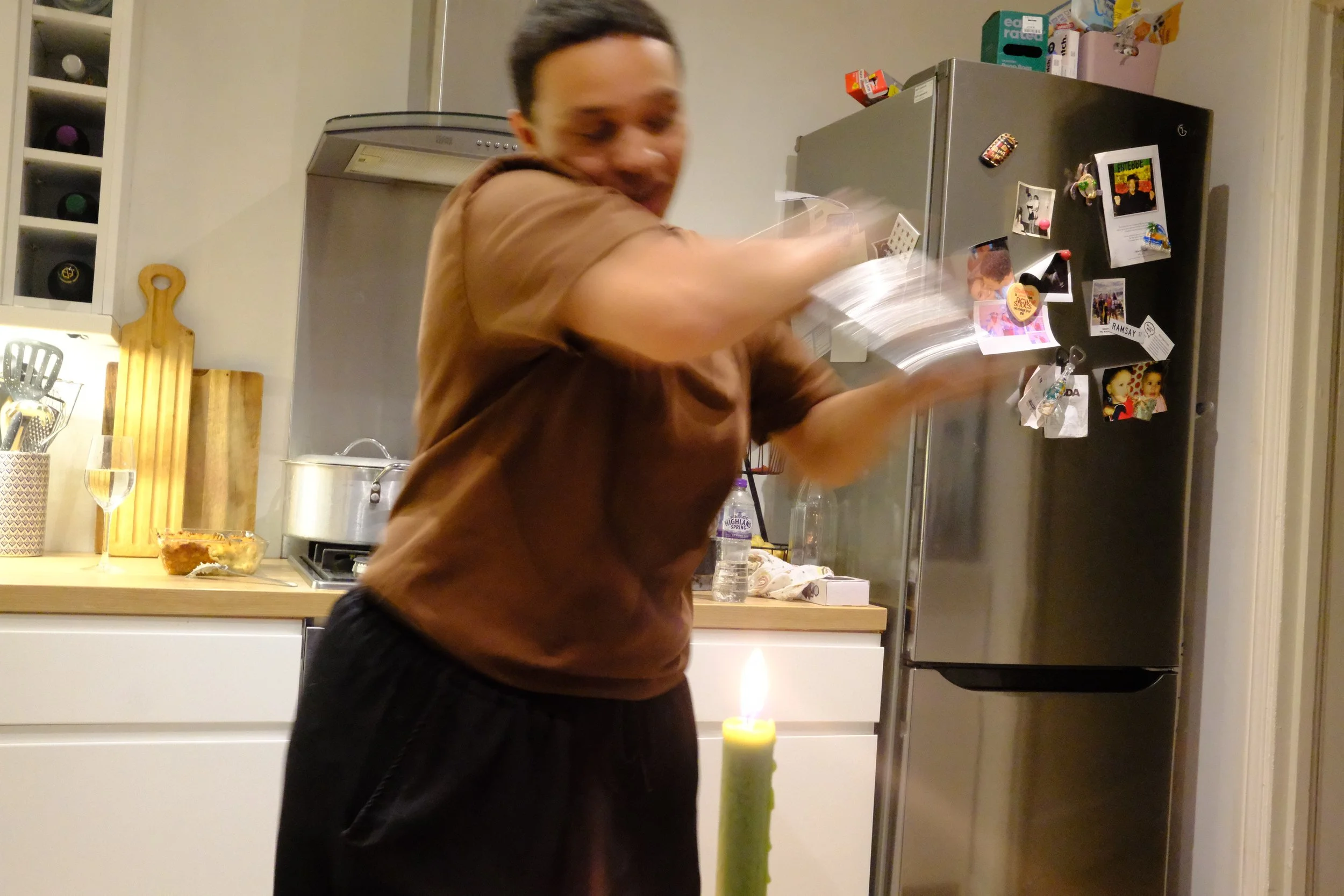 Person swinging a book in front of a refrigerator in a kitchen, with a lit candle in the foreground and various items on the countertop.