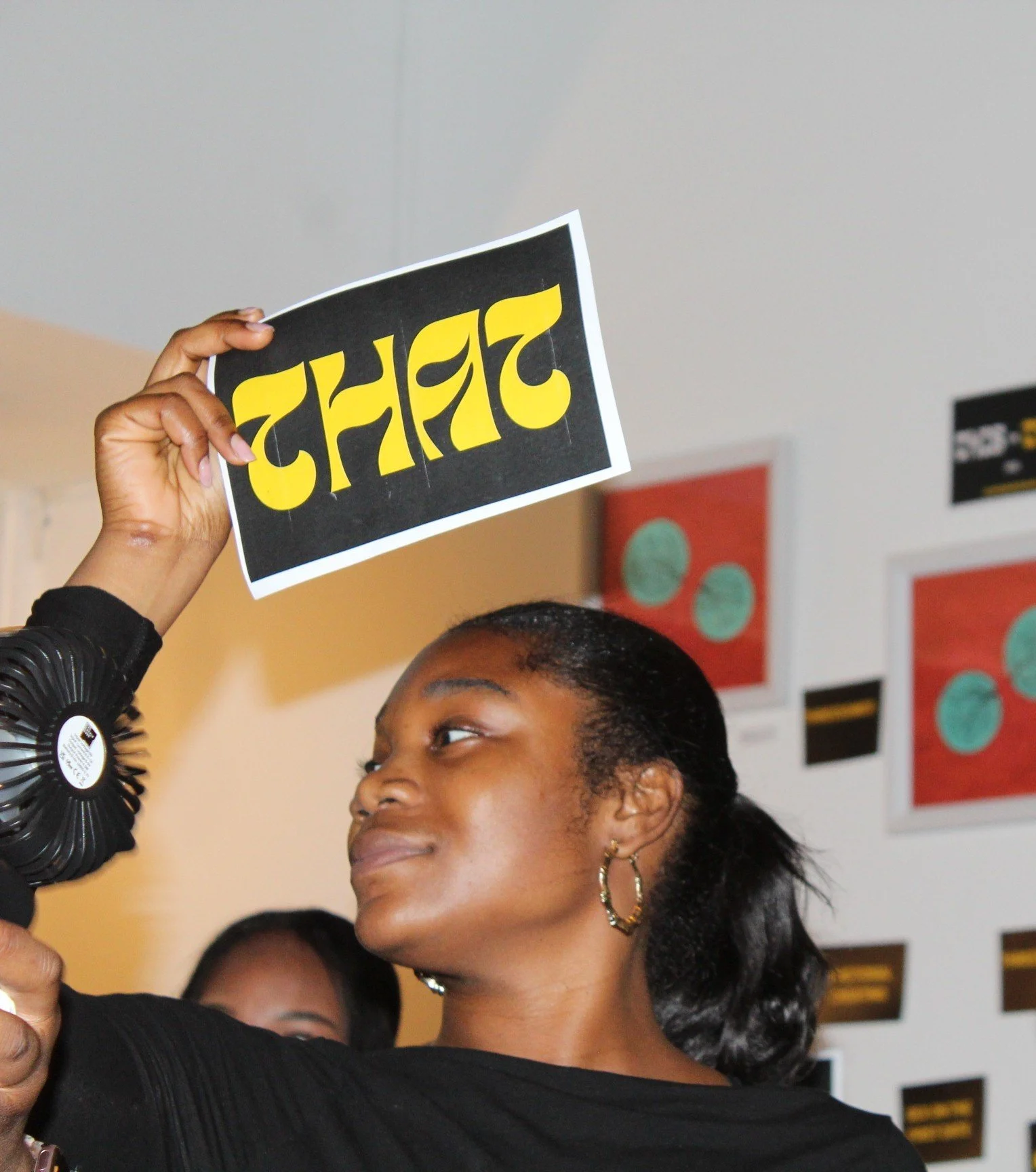 A woman holding a sign with the word 'Caught' written backwards, with a slight smile and a focused expression, in an indoor setting with colorful artwork on the wall.